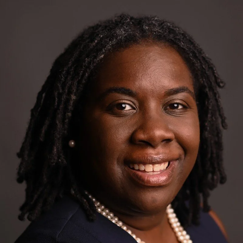 Headshot of an Belinda, wearing pearl jewelry, smiling against a dark background.