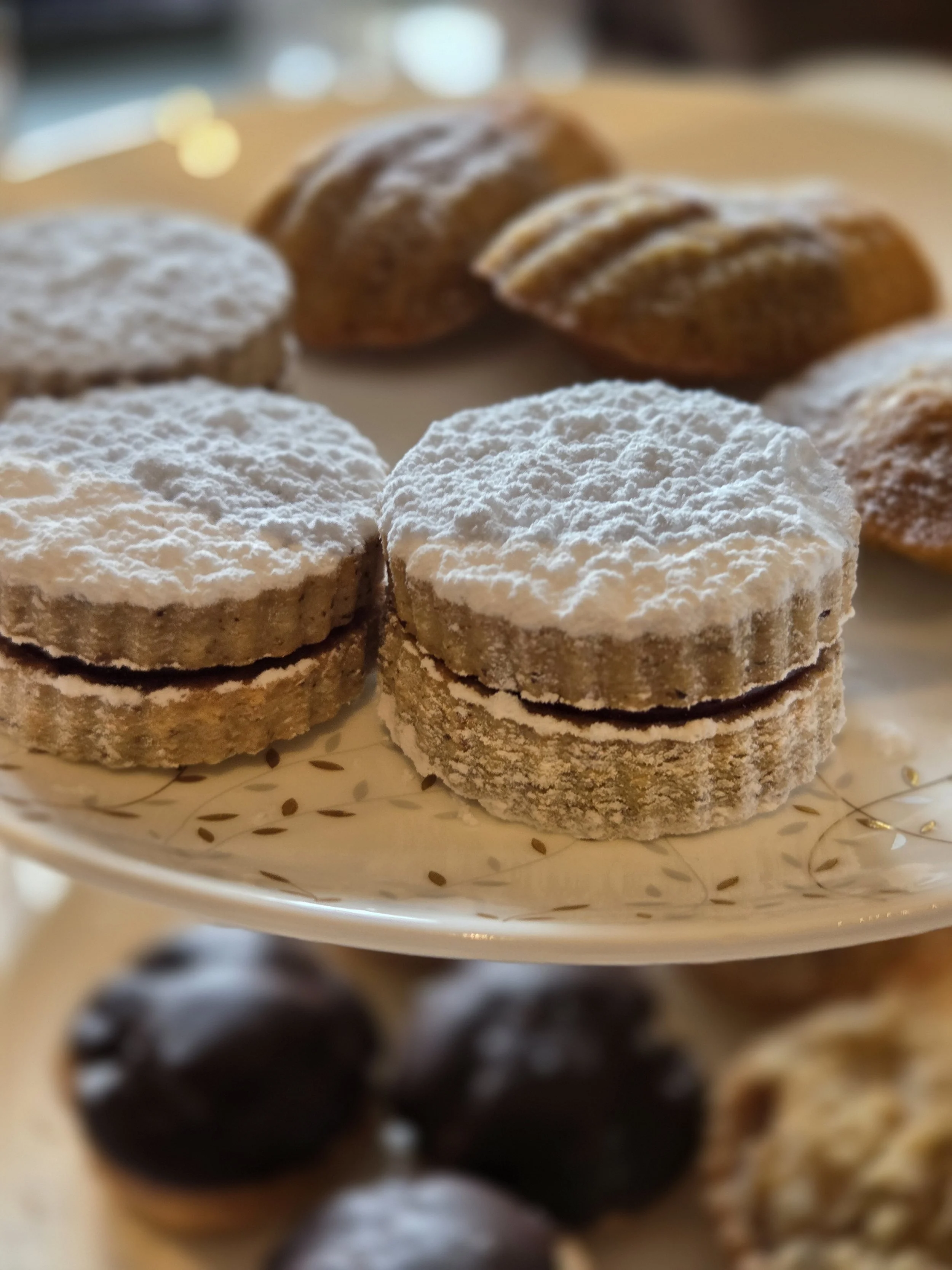 Plate of powdered sugar-dusted sandwich cookies with a dark filling, surrounded by assorted cookies on a decorative plate.