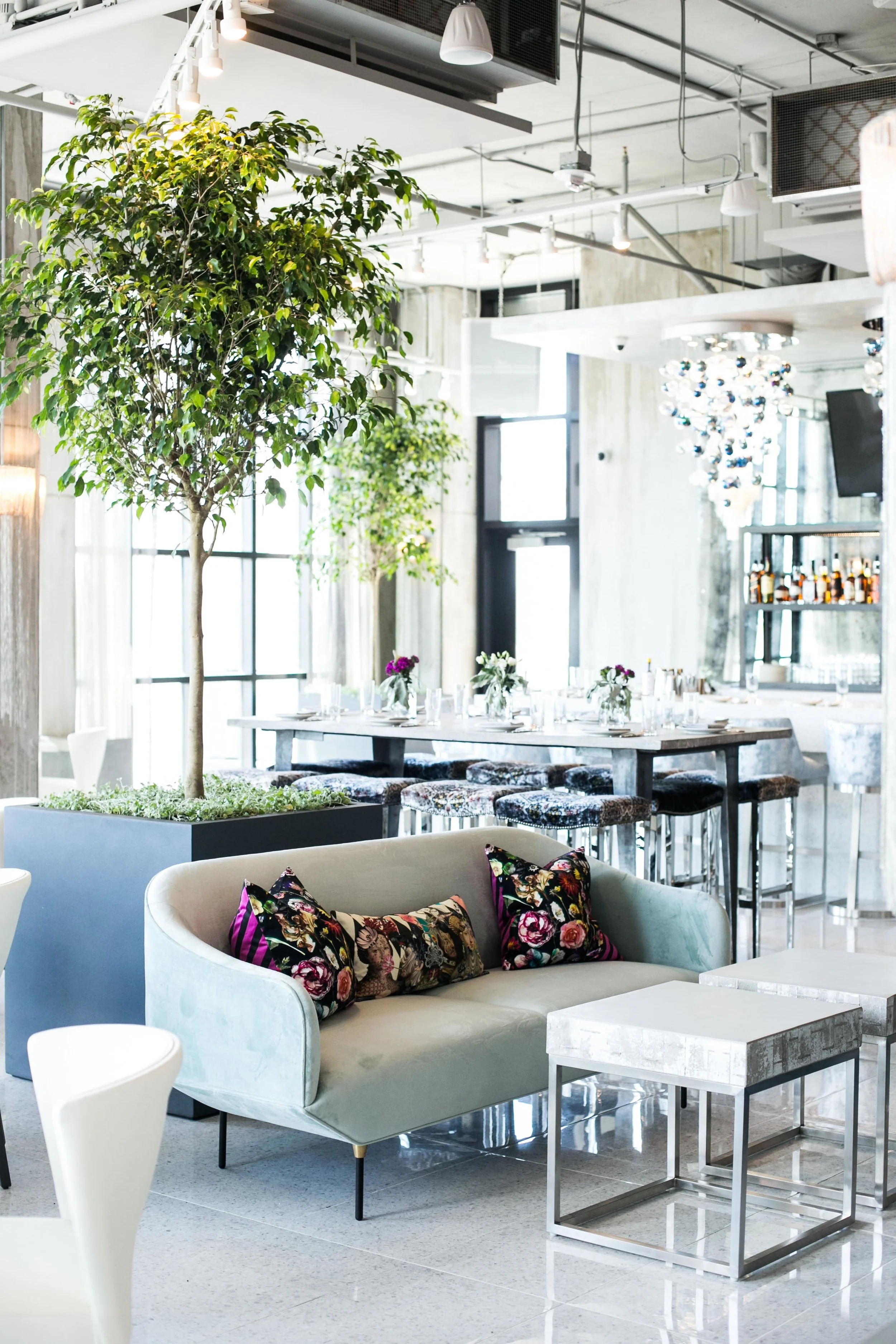 Modern restaurant interior with a light-colored sofa with floral pillows, potted trees, marble-top tables with floral centerpieces, bar stools, decorative hanging lights, and large windows letting in natural light.
