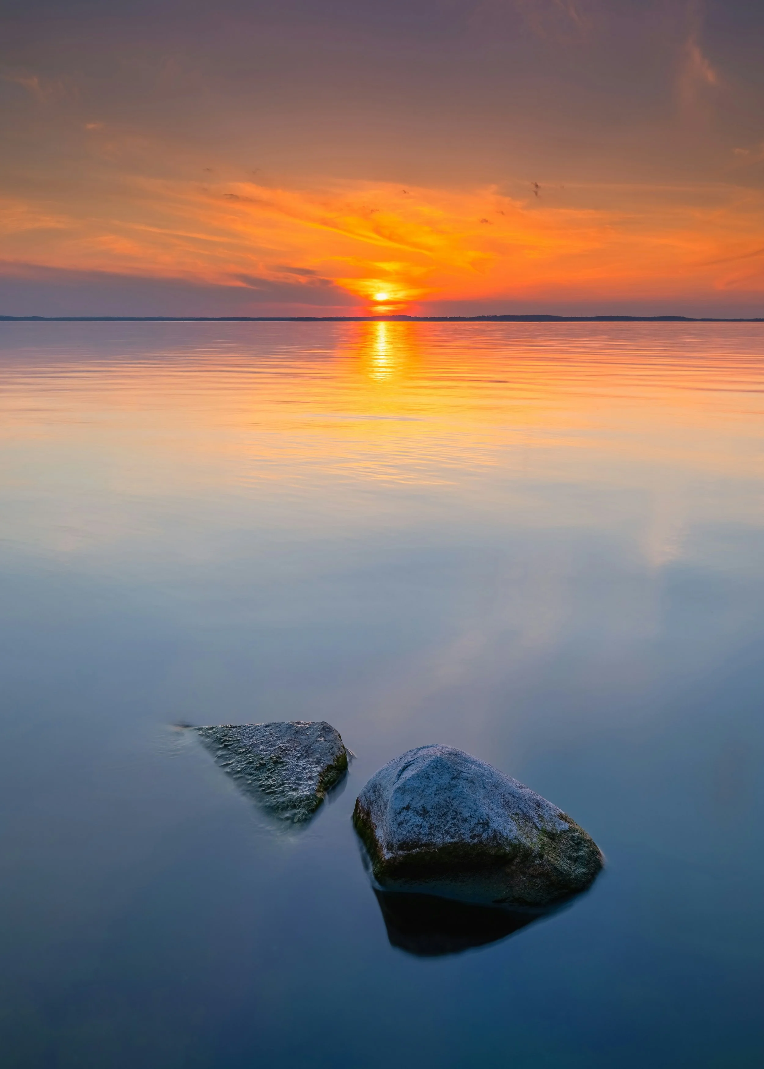 Sunset over calm water with a reflection of the sky and two rocks partially submerged in the foreground.