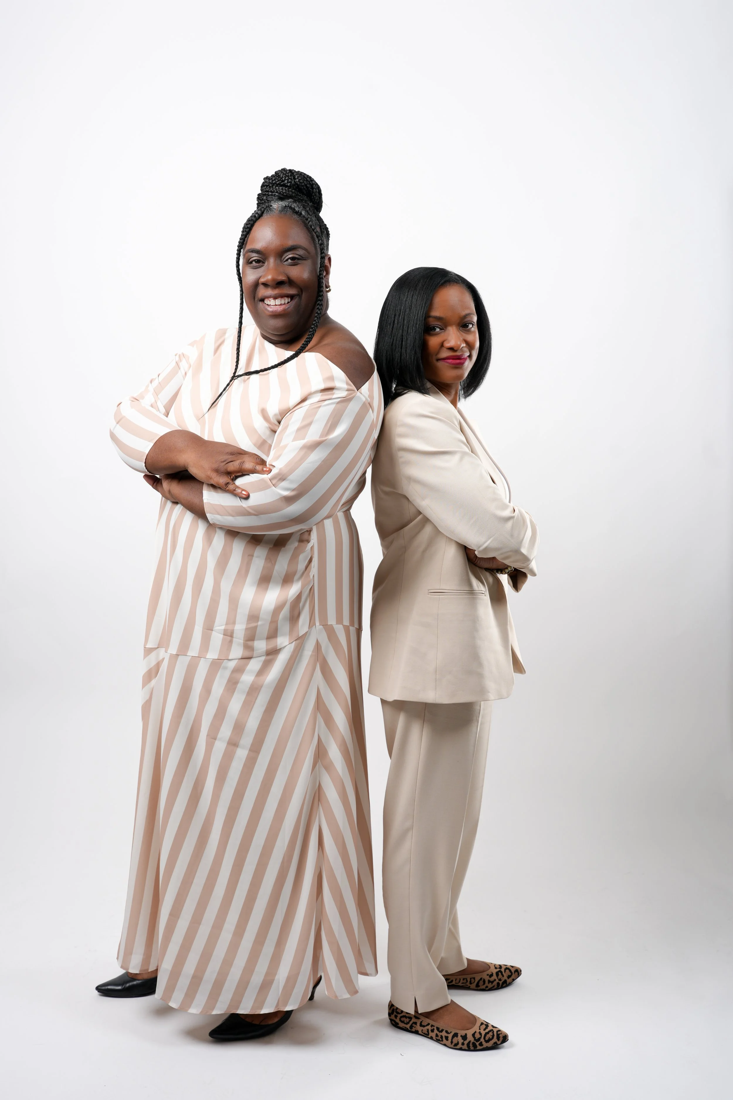 Two women standing back-to-back with arms crossed, smiling and looking at the camera, in a studio with a plain white background.