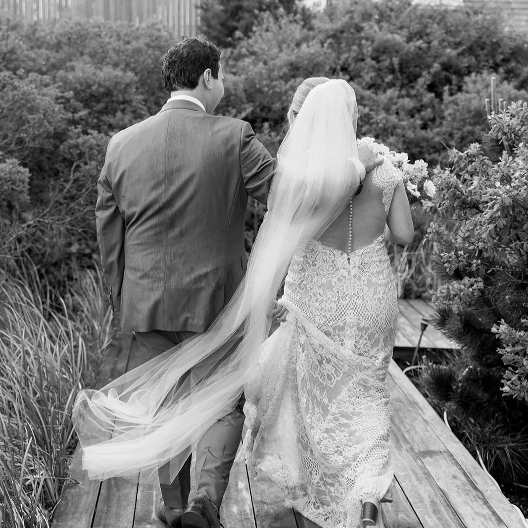 Wooden pathway leading towards the ocean, flanked by green bushes on both sides, with a partly cloudy sky overhead.