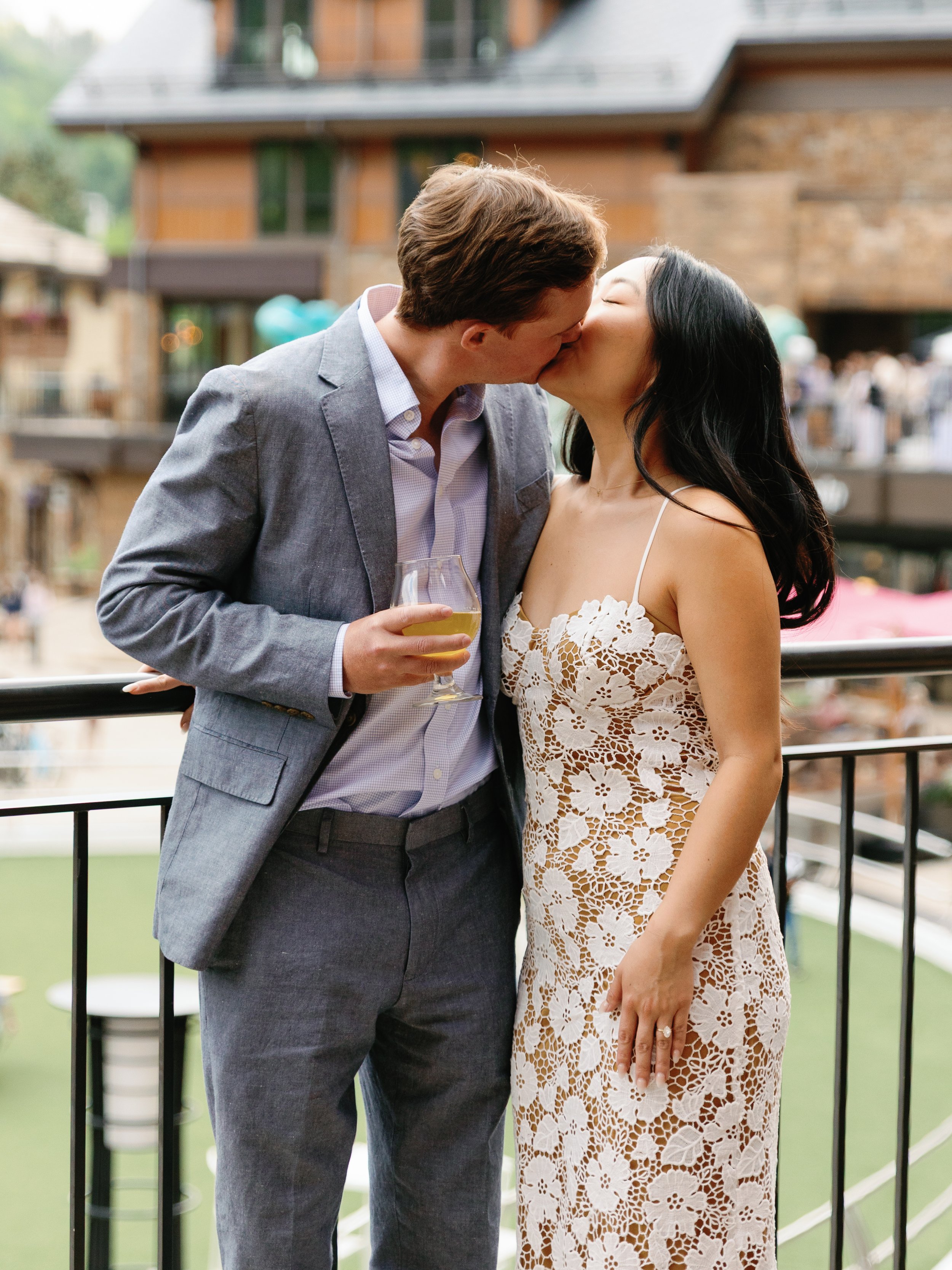 A couple kissing at an outdoor event, with the man holding a glass of white wine, on a balcony overlooking a gathering.