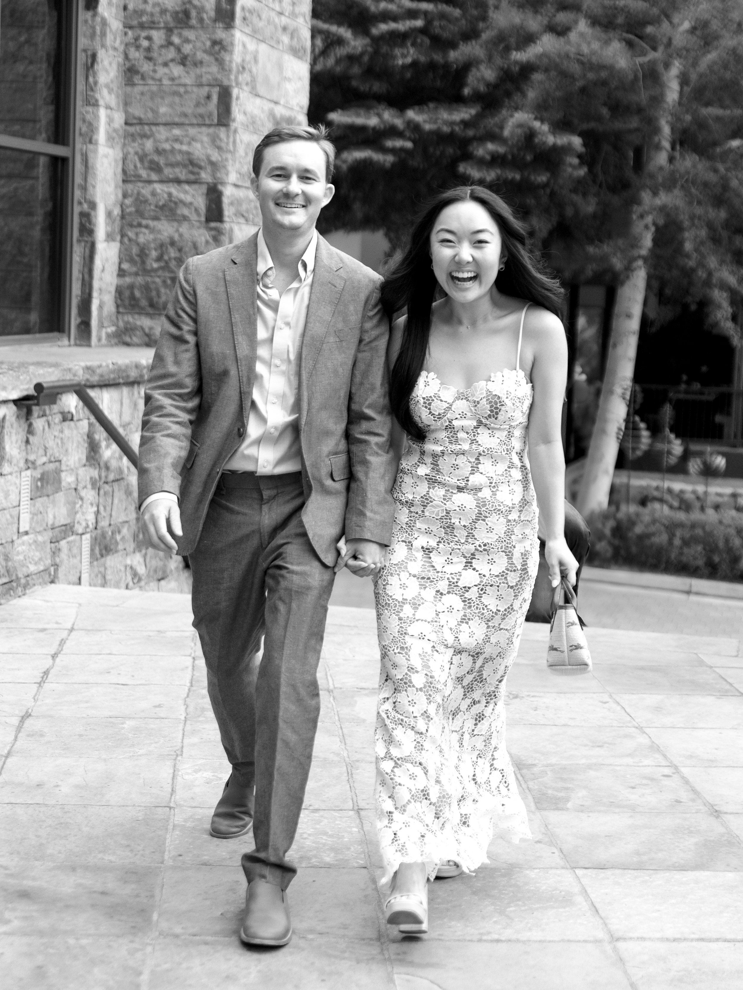 A smiling couple walking hand in hand outdoors, both dressed elegantly. The man wears a suit jacket and pants, and the woman wears a lace dress. They seem joyful and are walking on a paved pathway with trees and a stone building in the background.