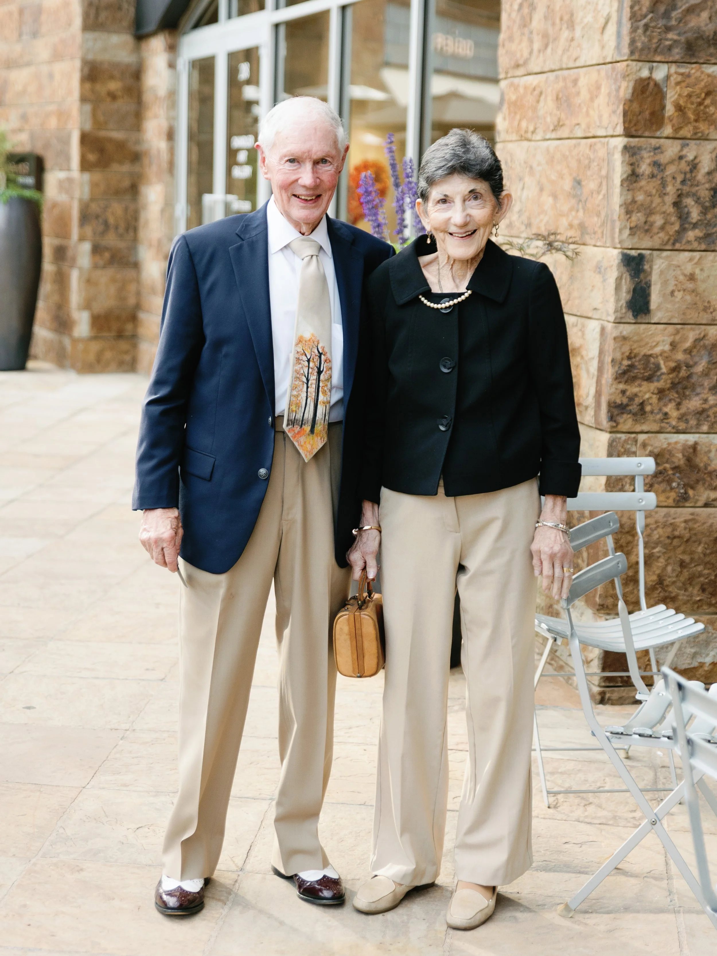 An elderly man and woman standing outside on a paved area, smiling, dressed in formal attire. The man is wearing a navy blazer, white dress shirt, patterned tie, and beige trousers, while the woman is wearing a black jacket, beige trousers, and holdi