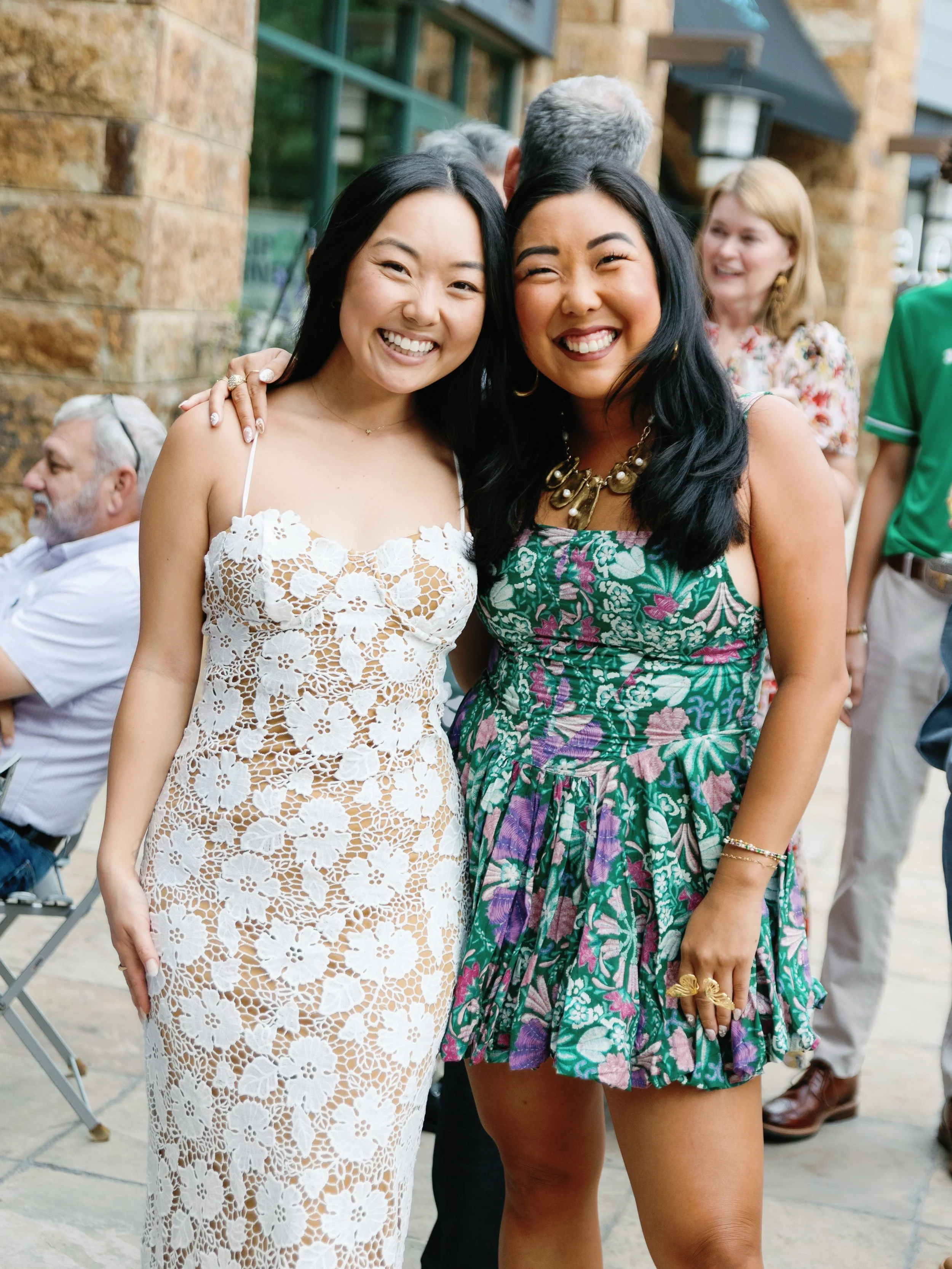 Two women smiling and standing close together outdoors at a social gathering, with other people in the background.
