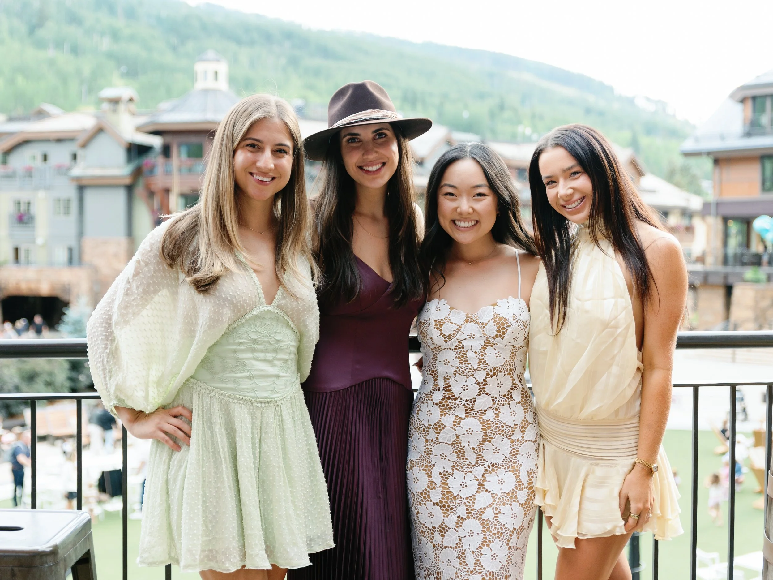 Four women standing on a balcony outdoors, smiling at the camera, with a mountain and village in the background.