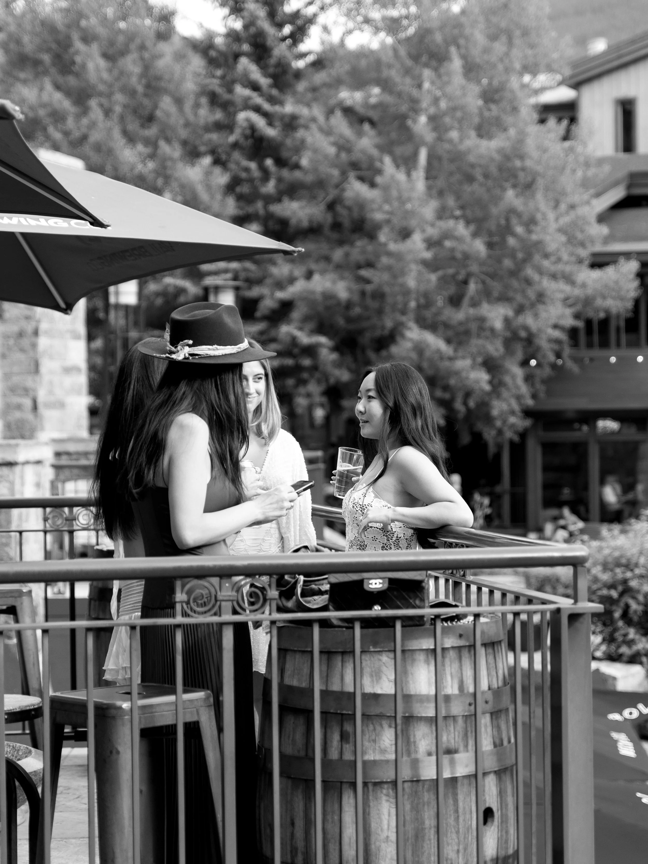Three women having a conversation on an outdoor patio or balcony, one holding a glass of drink, with trees and buildings in the background.
