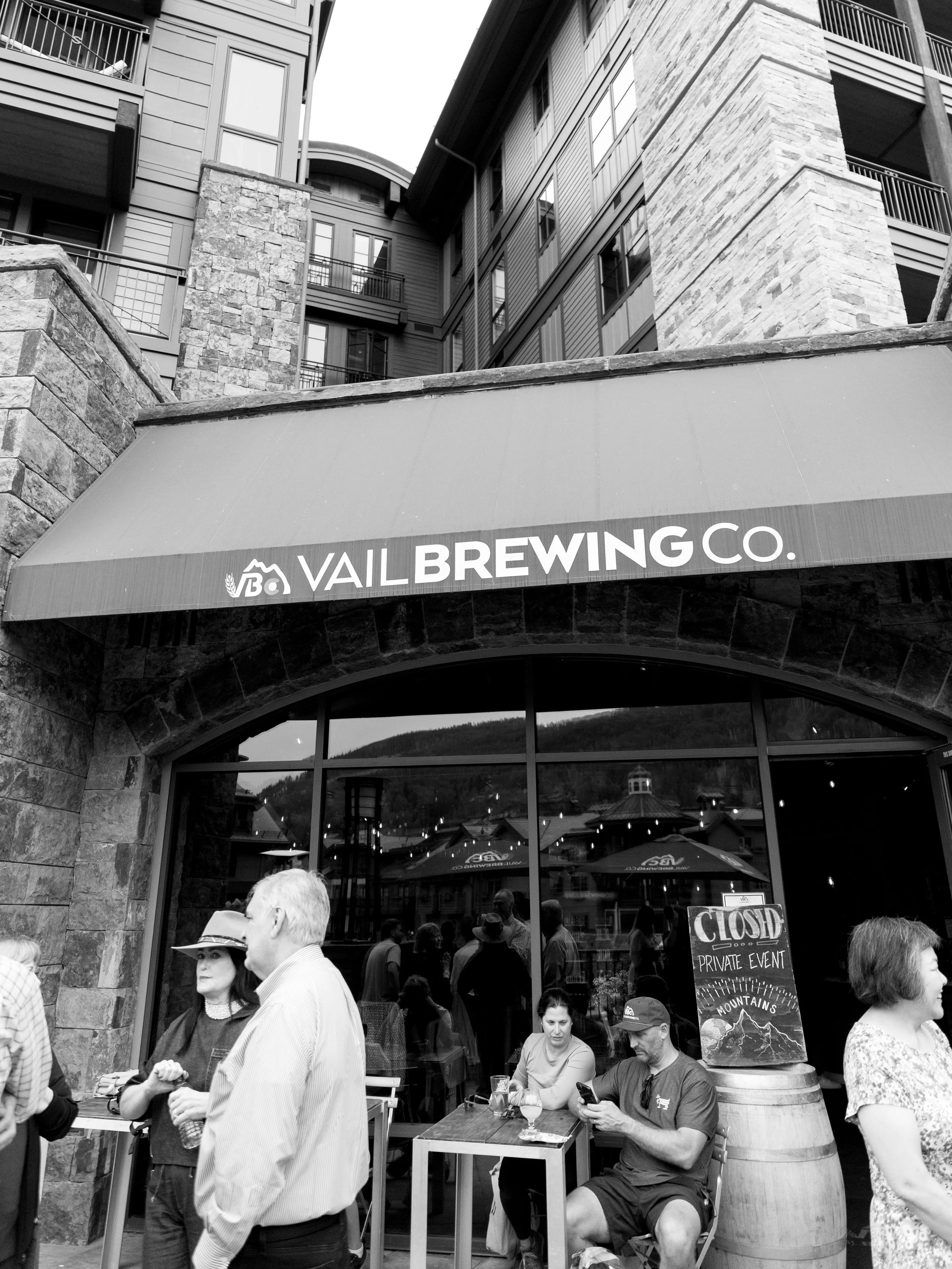 People socializing outside a bar with a sign that reads 'CLOSED, Private Event, Mountains' in front of Vail Brewing Co., with a building and mountains reflected in the large window.