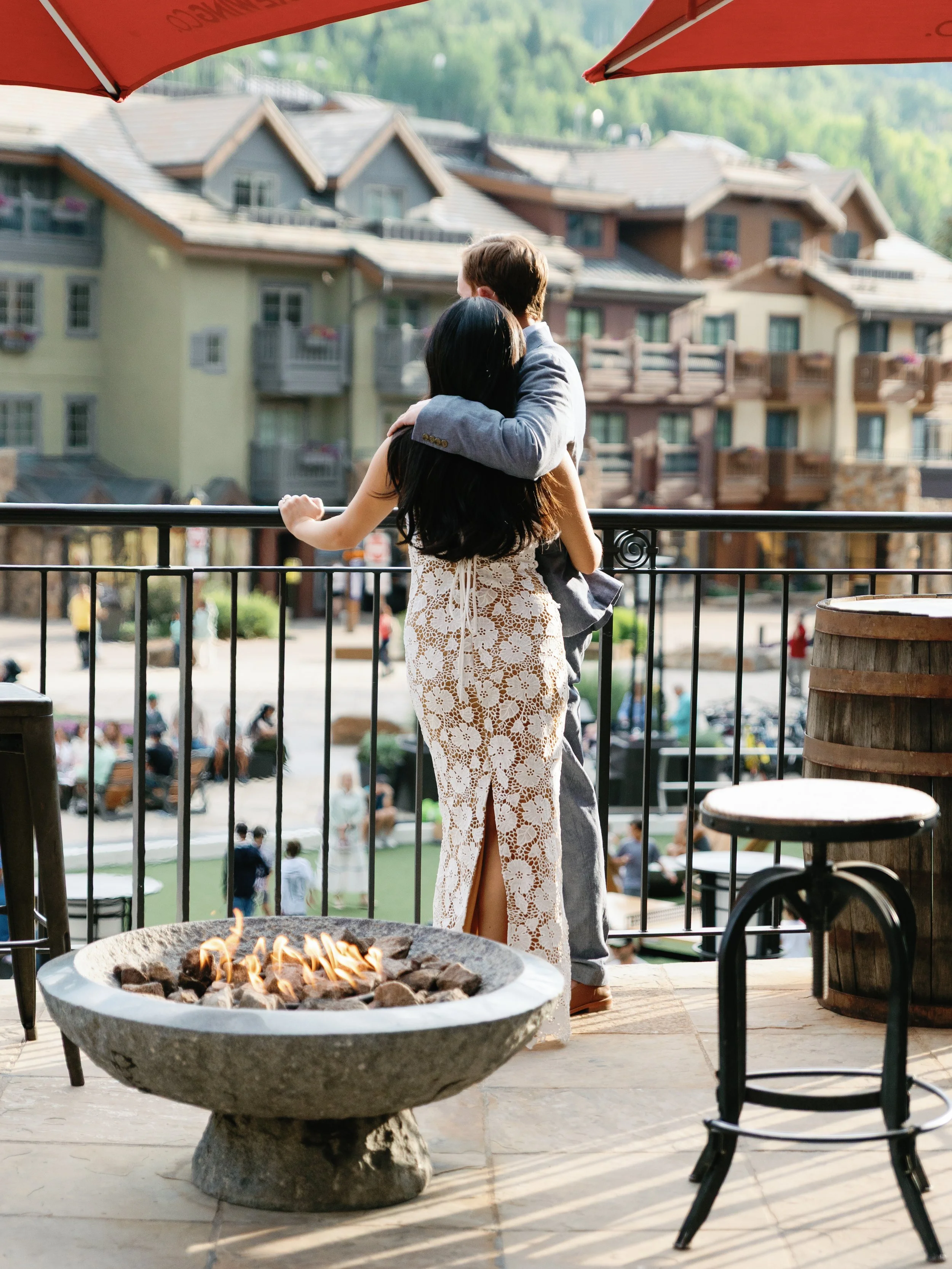 A couple embracing on a balcony overlooking a busy outdoor area with buildings in the background. There is a fire pit on the patio.