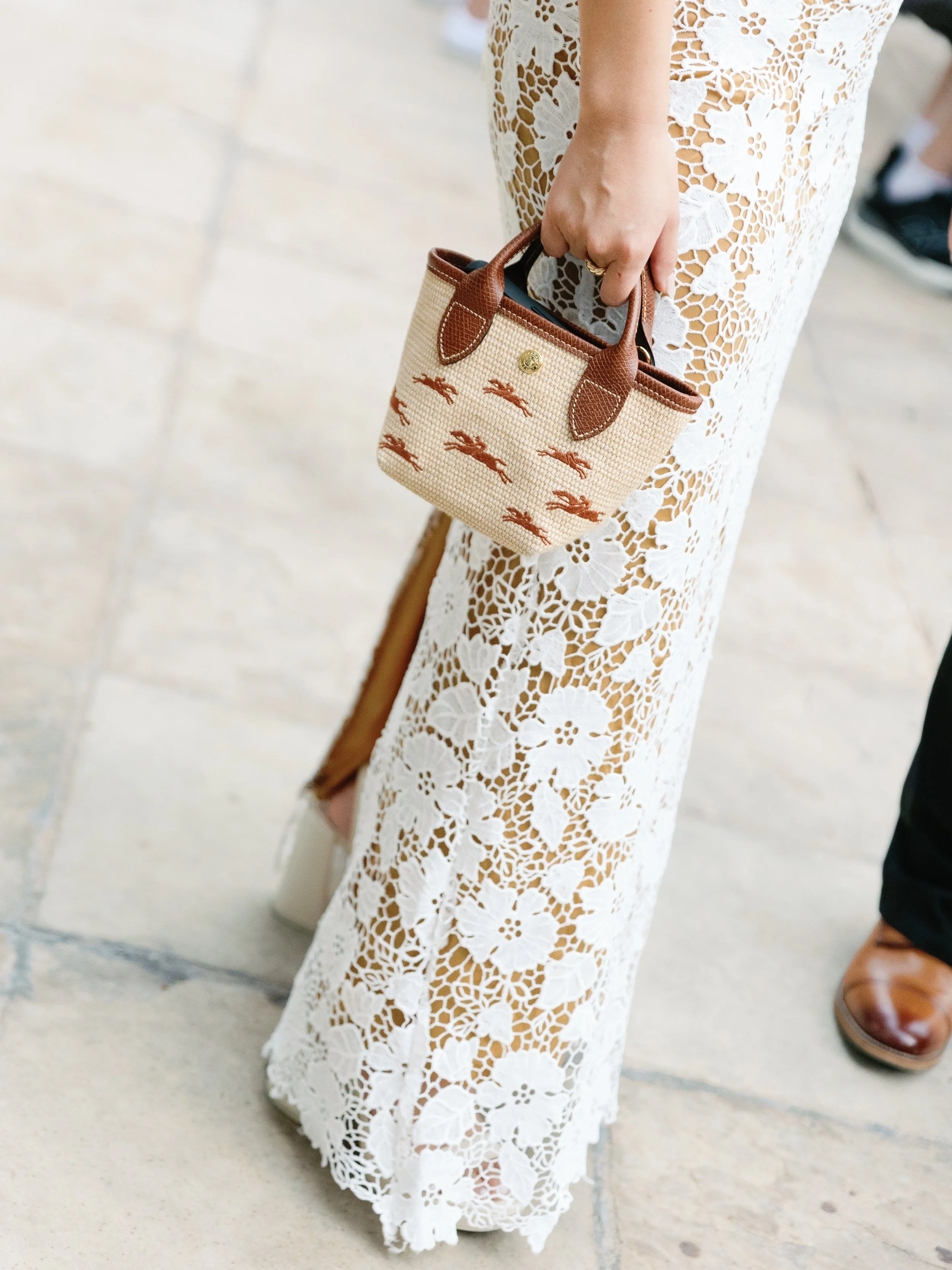 A woman wearing a white lace dress holding a small beige and brown handbag with a pattern of brown animals, standing on tiled flooring.