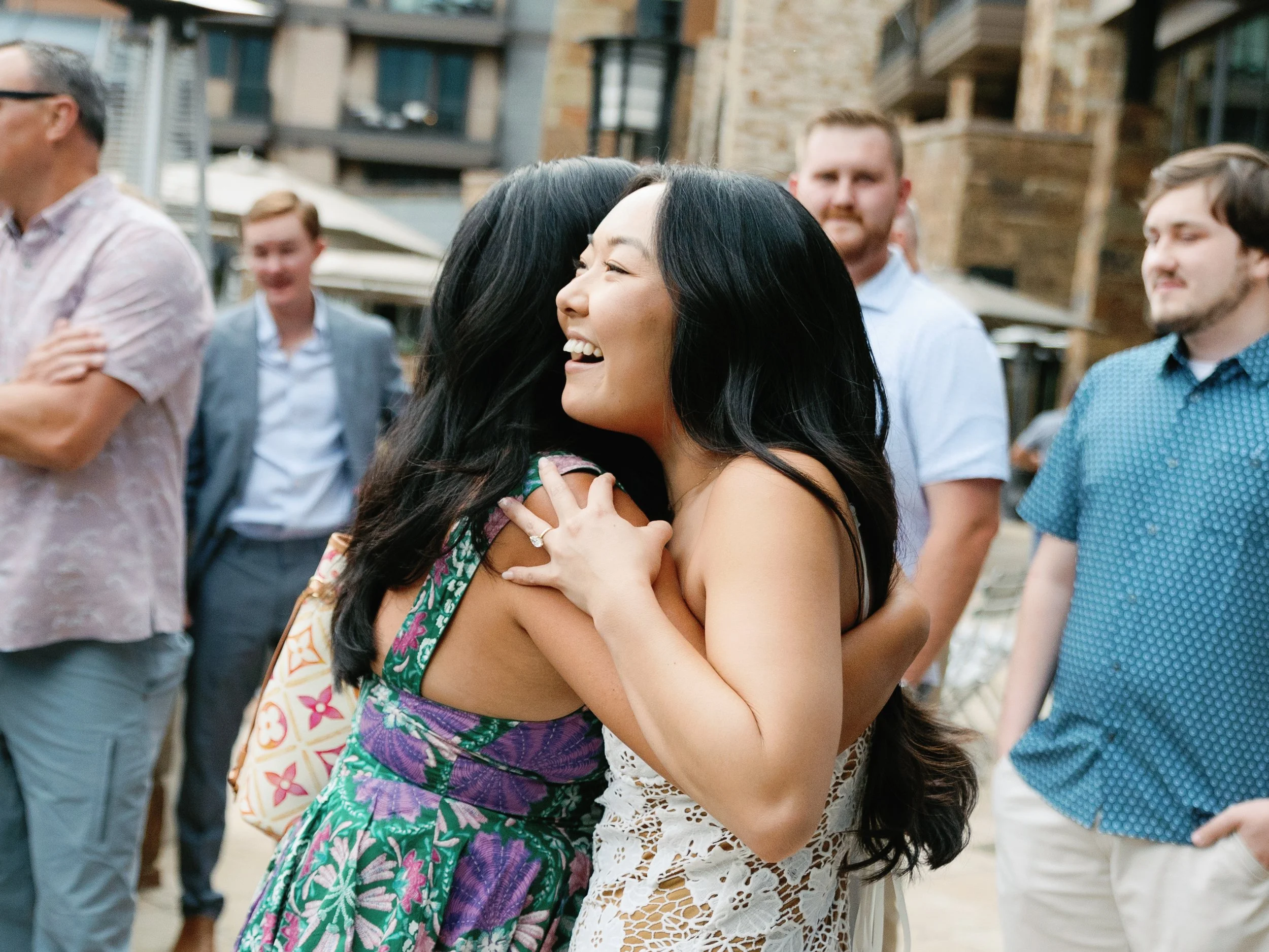Two women hugging and smiling at an outdoor social gathering, with several onlookers in casual attire in the background.
