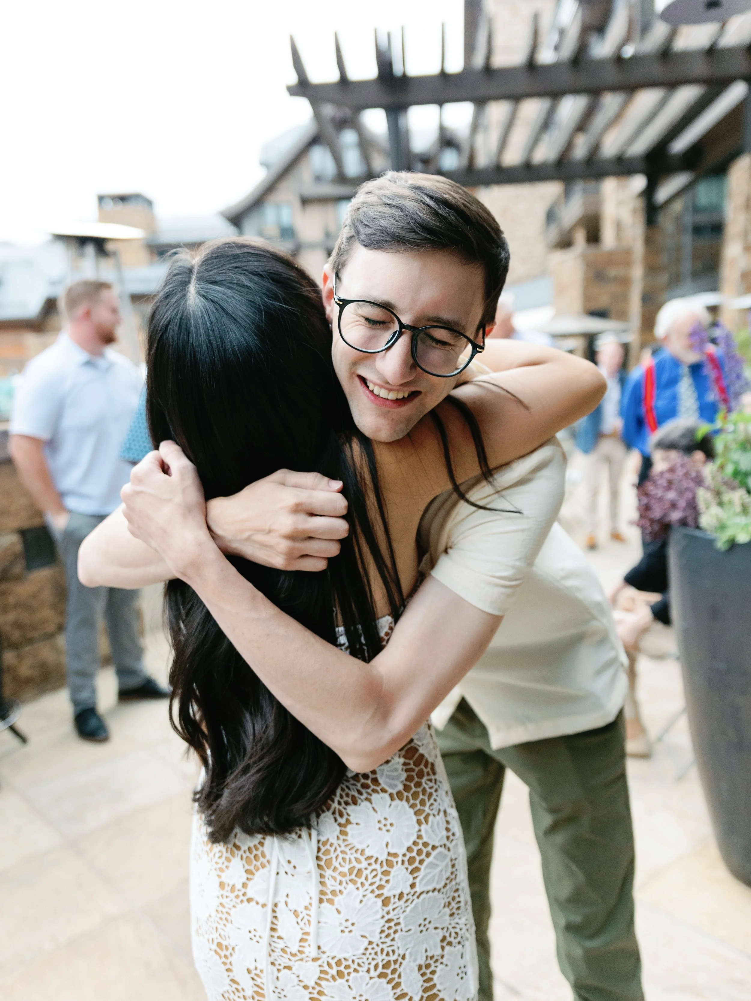 A young man with glasses is hugging a woman with long dark hair at an outdoor gathering. The woman is wearing a white lace dress, and the man is smiling with his eyes closed.