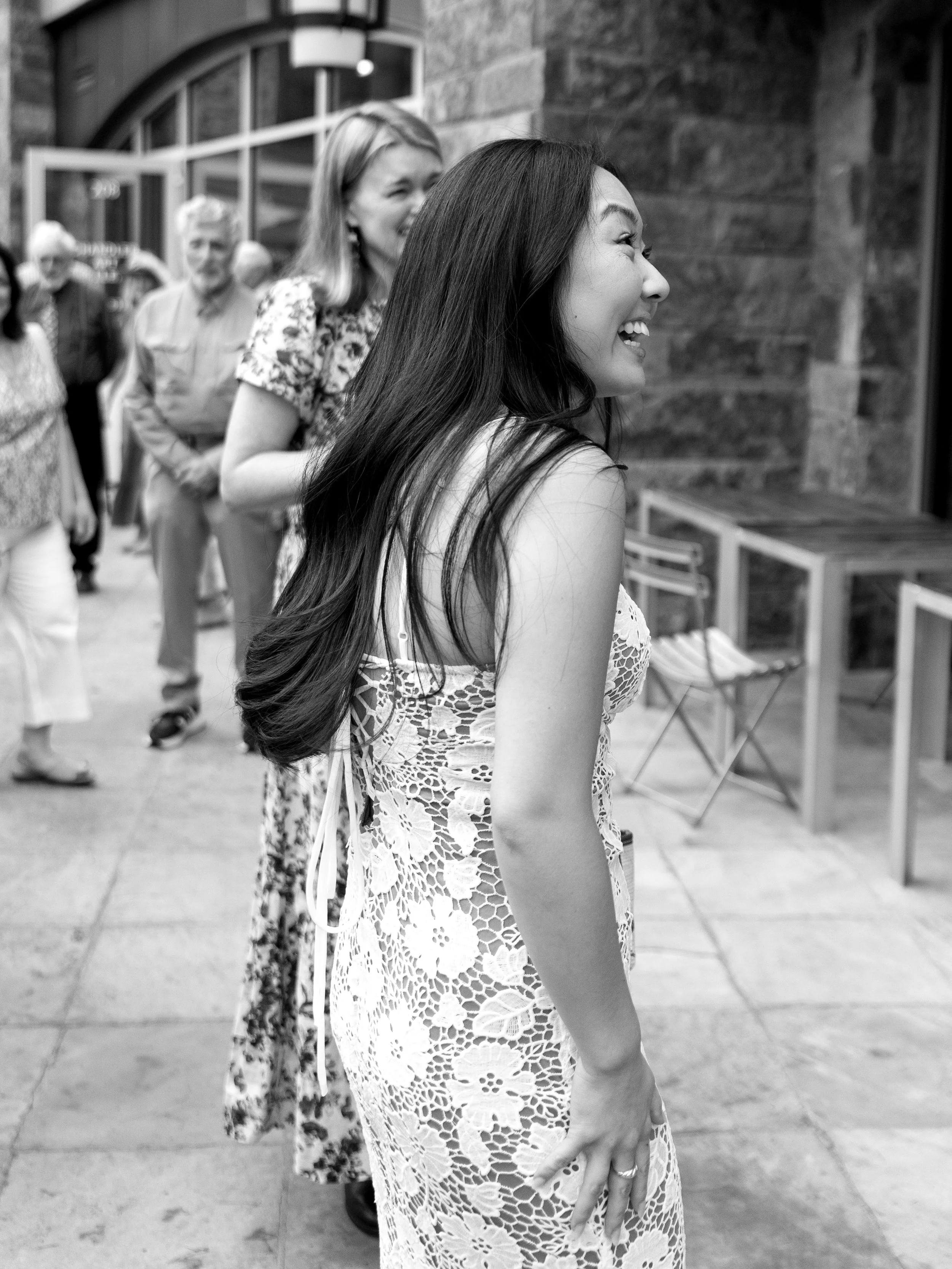 A woman with long dark hair laughing and smiling, wearing a sleeveless lace dress, standing outside near a brick wall with other people in the background.