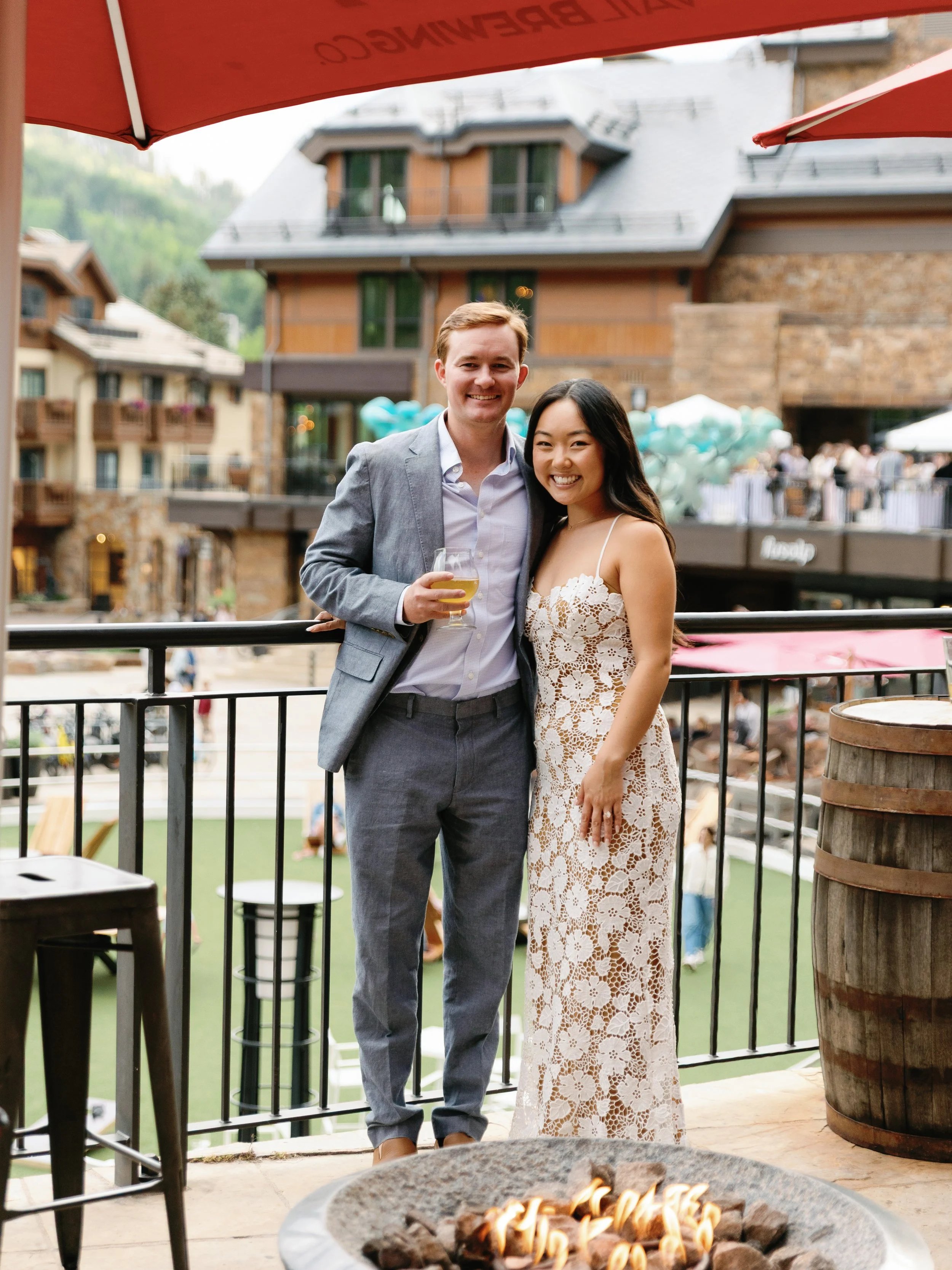 A couple stands on a balcony at an outdoor event, smiling and holding drinks. The man wears a light grey suit with a white shirt, and the woman wears a white lace dress. There are balloons and people in the background.