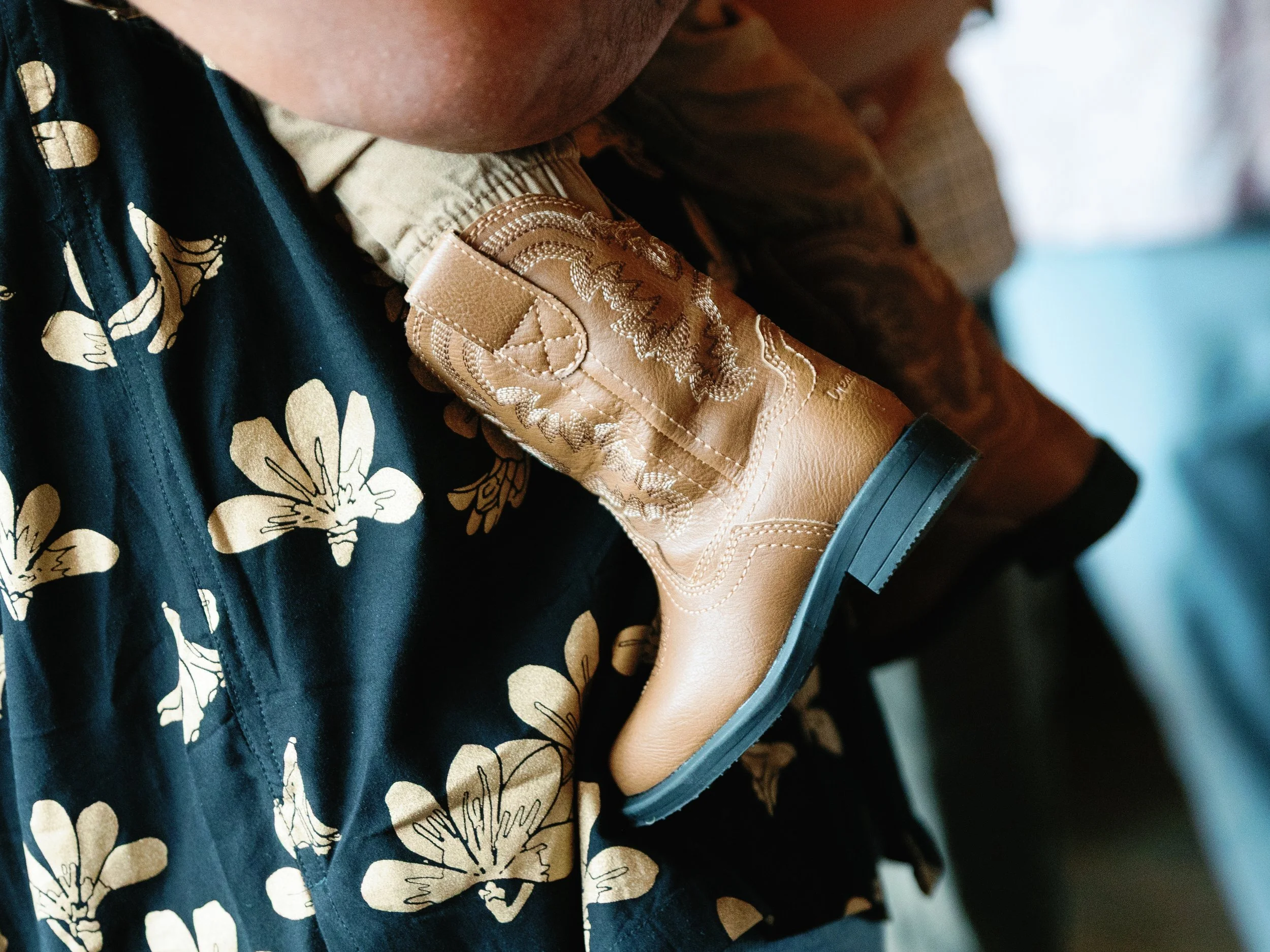 A person in cowboy boots leaning against a floral patterned shirt, with a background of blurred indoor setting.