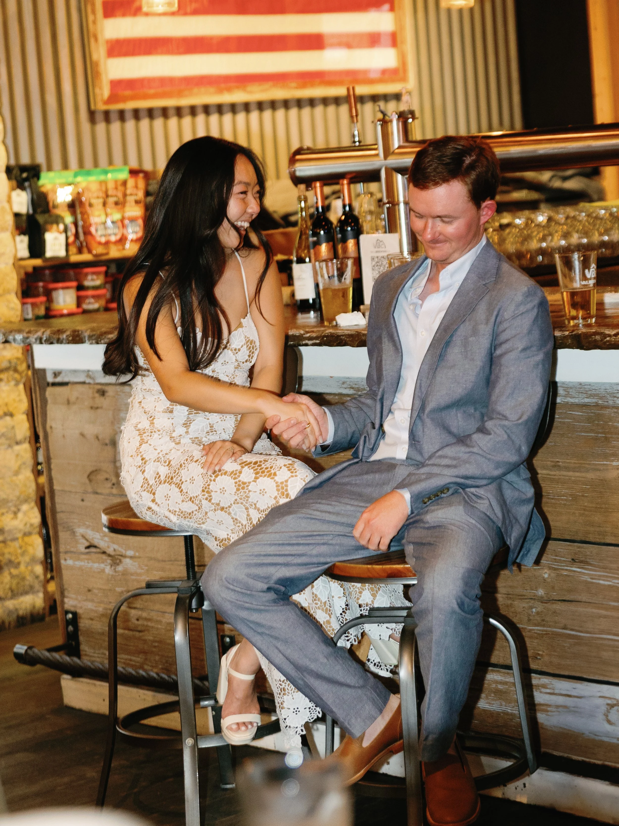 A woman and a man in formal attire are sitting on barstools at a bar, holding hands and smiling. The woman is wearing a white lace dress, and the man is wearing a light gray suit. There are bottled drinks and glasses of beer on the bar counter behind