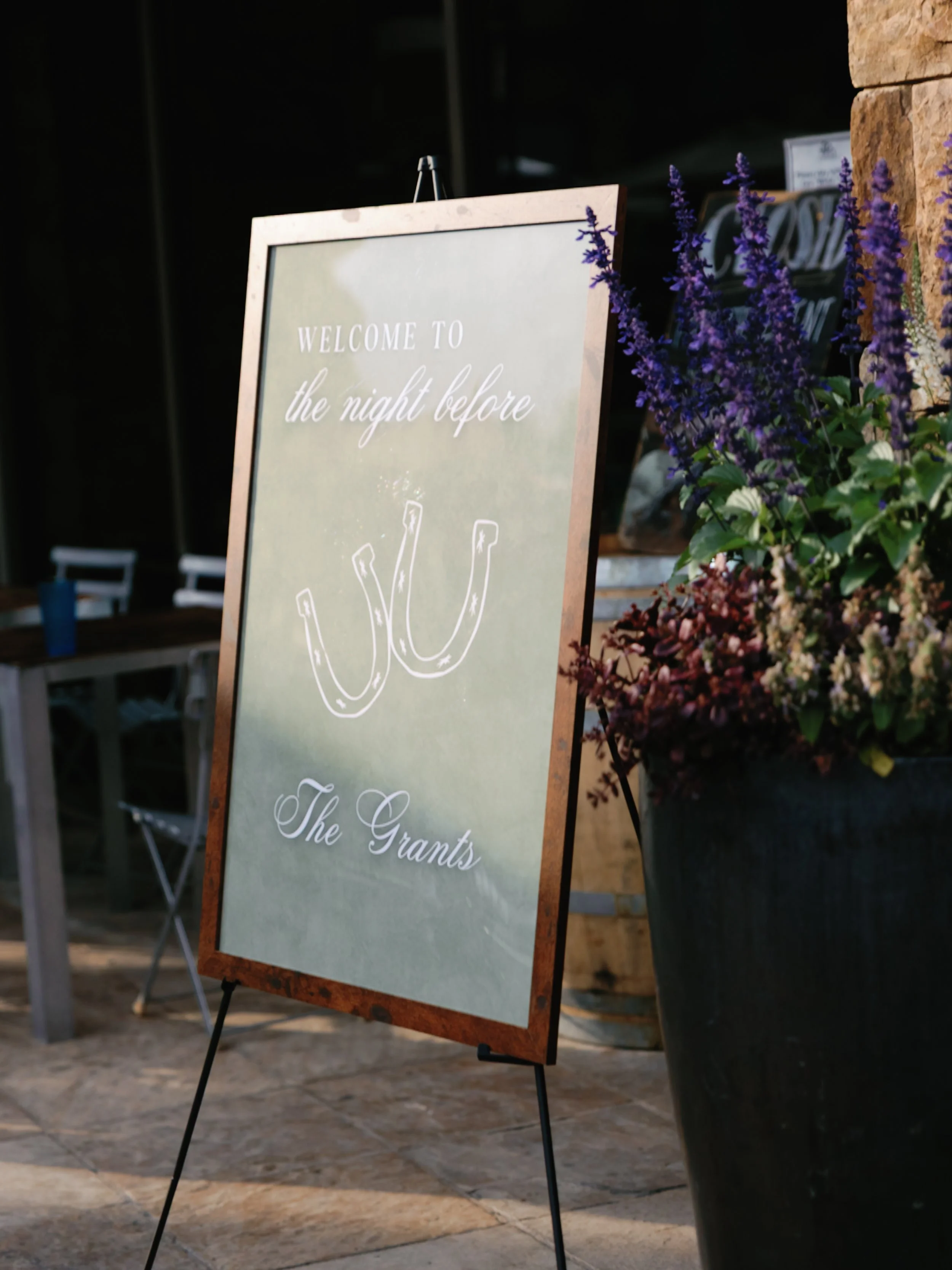 A signboard reading 'Welcome to the night before the Grants' with a horseshoe illustration, placed outside near purple flowers.
