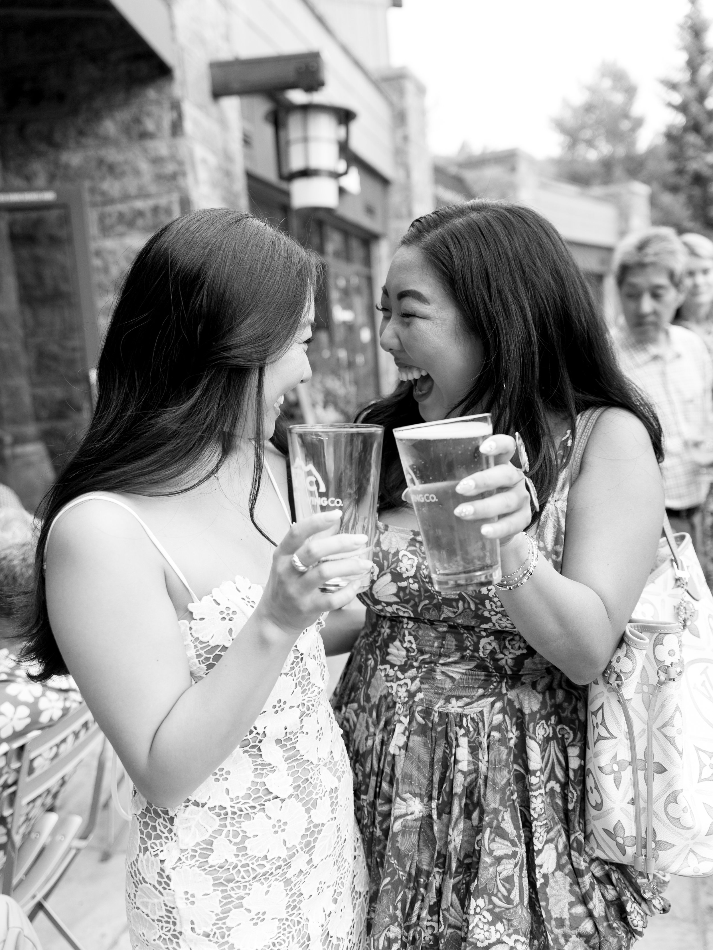 Two women enjoying drinks and laughing together outdoors, with several other people and buildings in the background.