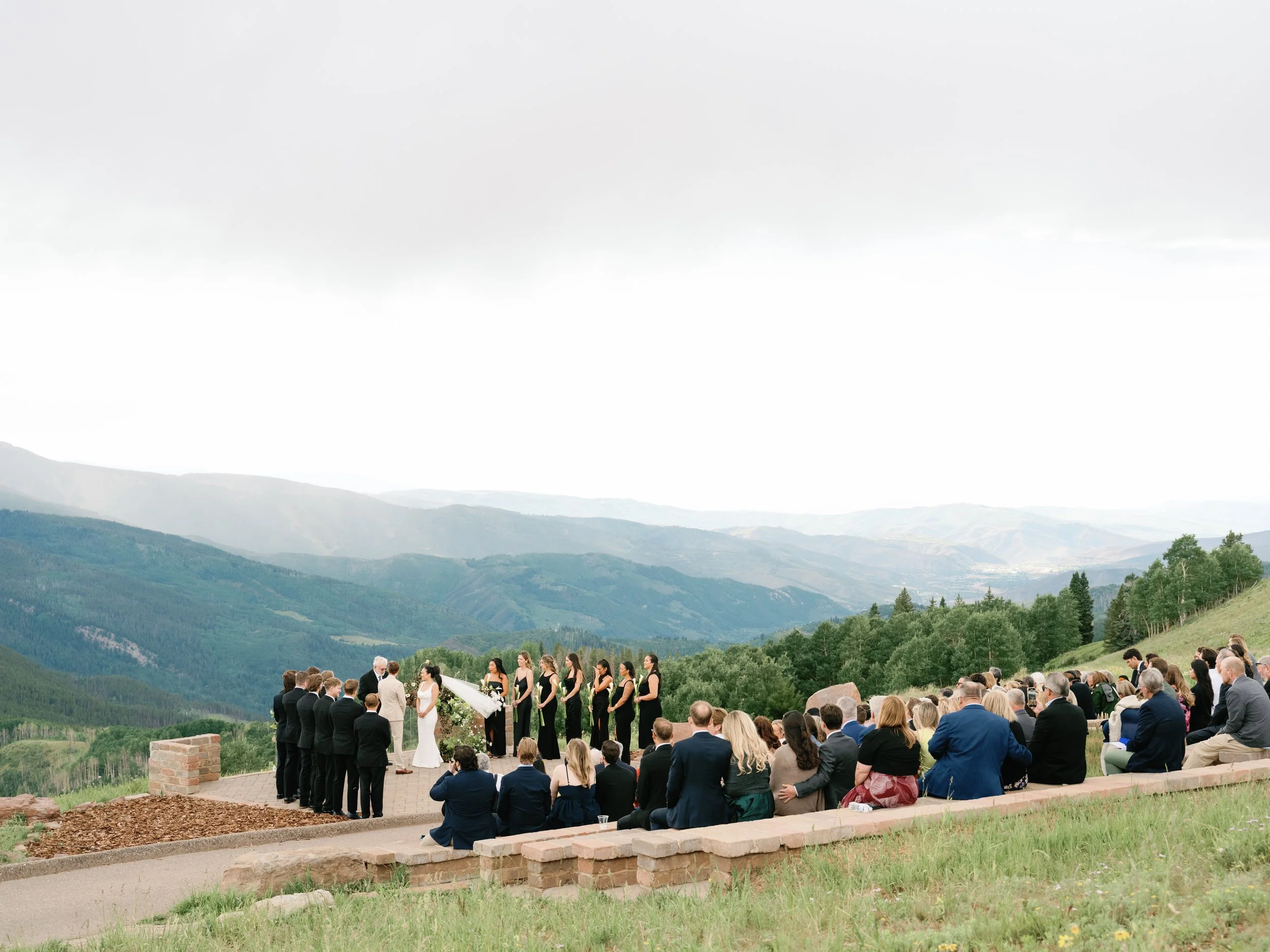 A wedding ceremony outdoors on a mountain overlook, with the bride and groom facing each other, surrounded by bridesmaids and groomsmen, with wedding guests seated on a hillside overlooking mountains.