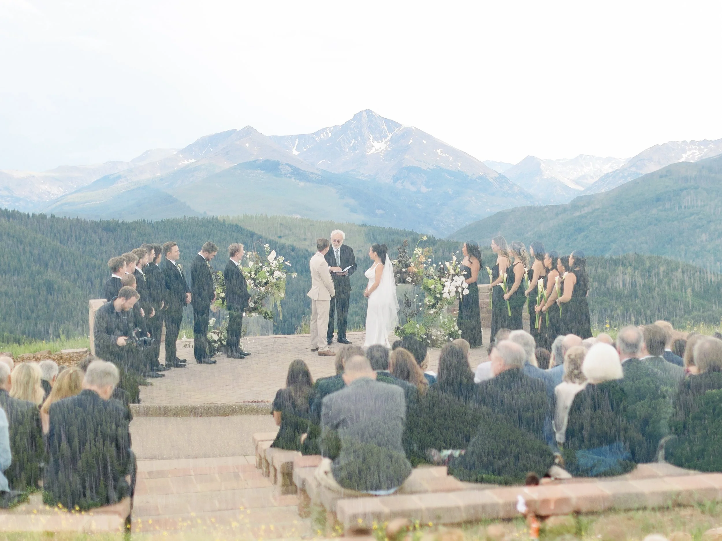 A mountain wedding ceremony outdoors with mountains in the background. The bride and groom stand before an officiant, surrounded by bridesmaids and groomsmen, while guests sit watching.