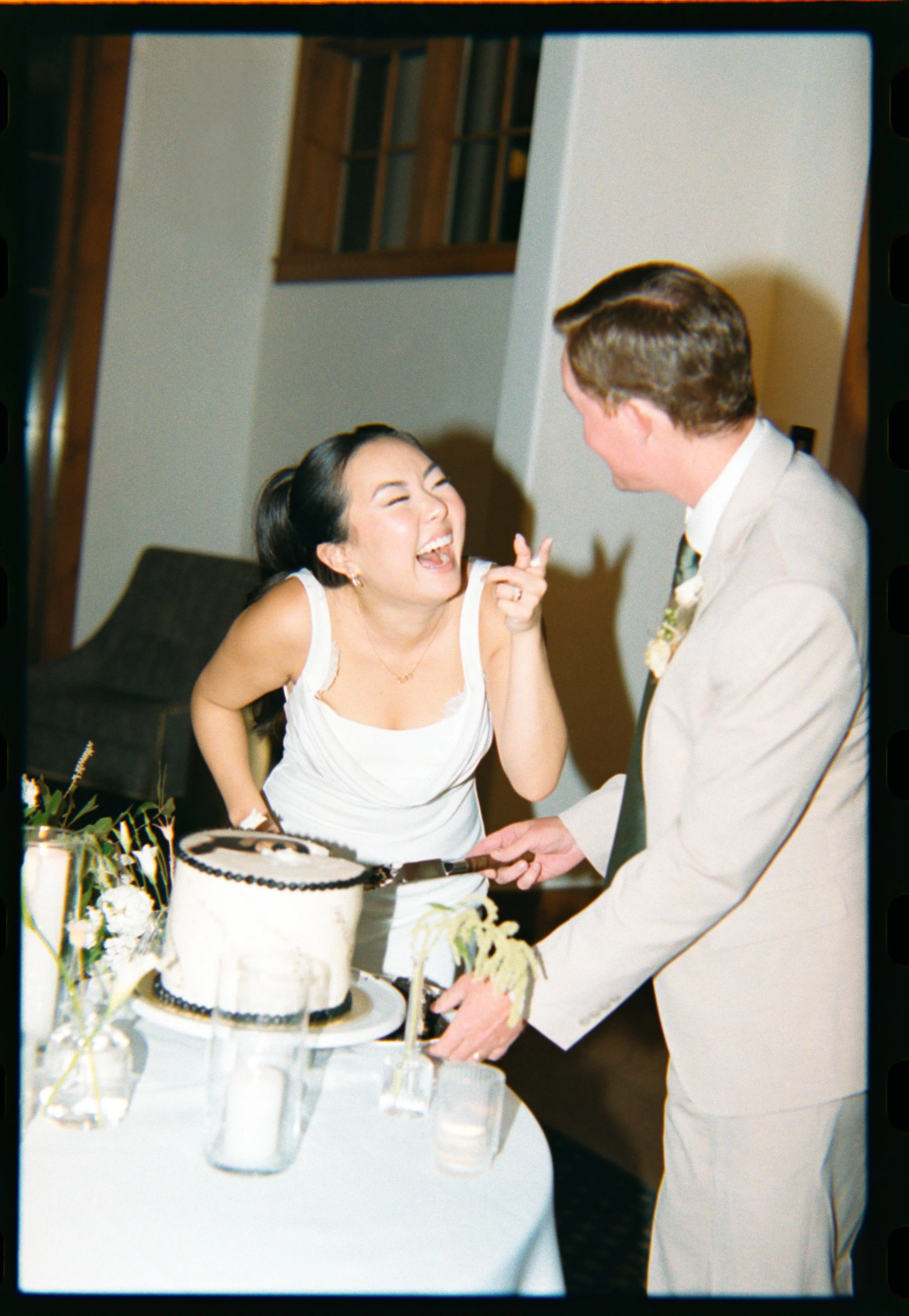 A woman in a white dress laughing and pointing at a man in a beige suit, who is holding a cake knife near a wedding cake, in a celebration setting.