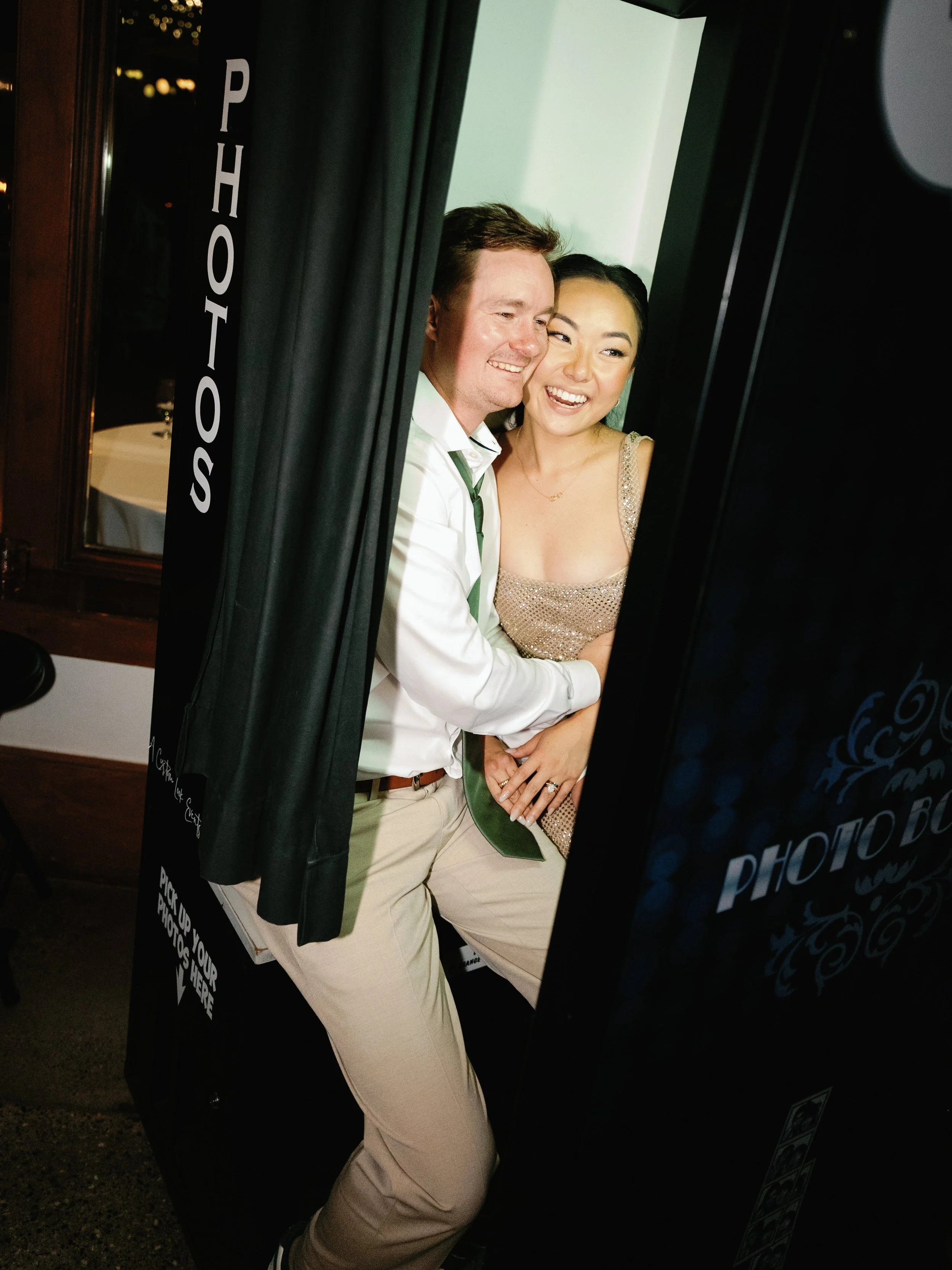 A couple smiling and posing in a photo booth with a 'Photos' sign on the side, black curtains, and a wooden frame.
