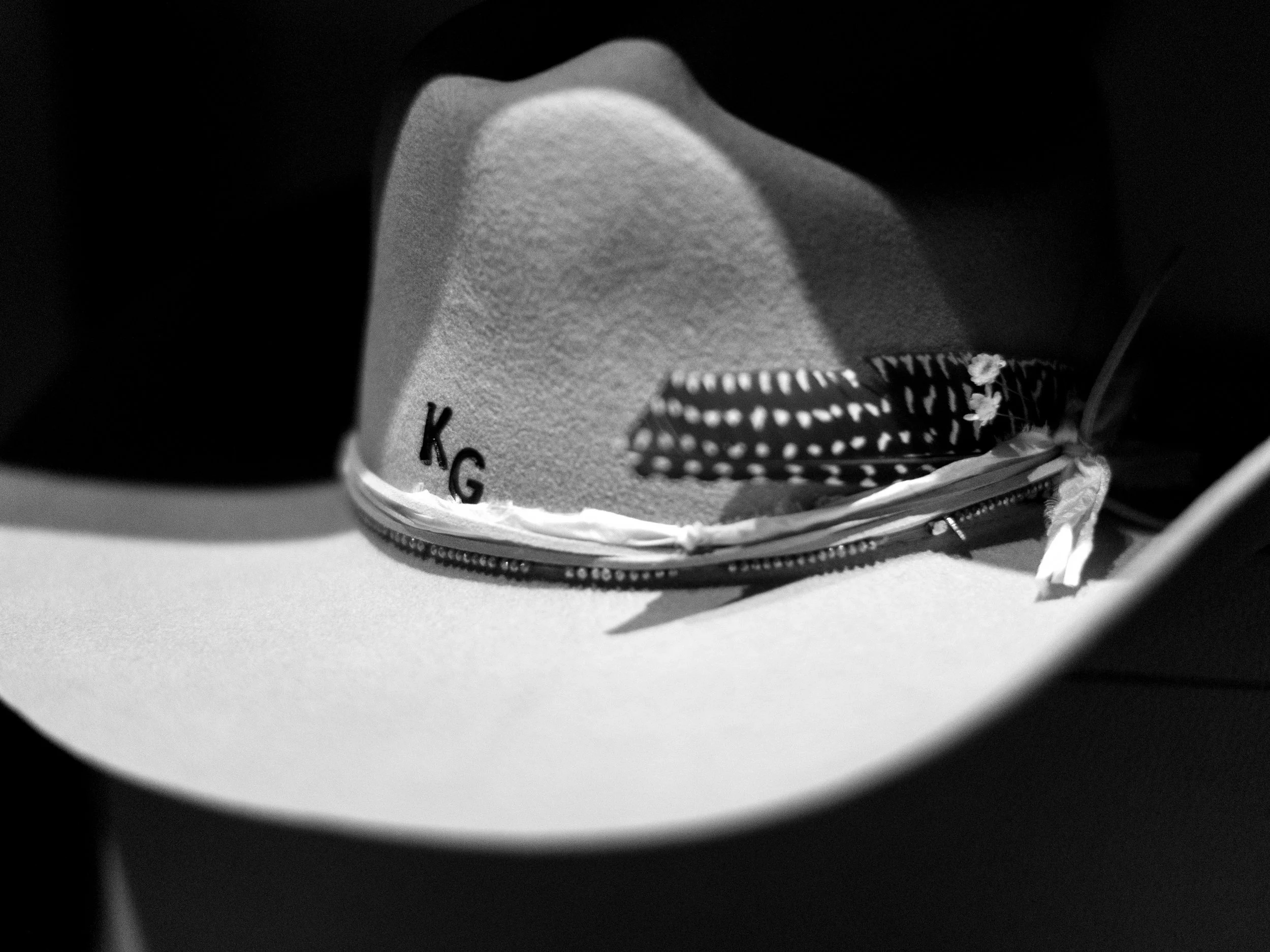 A cowboy hat with decorative band featuring a feather and the initials 'KG' embroidered on the side.