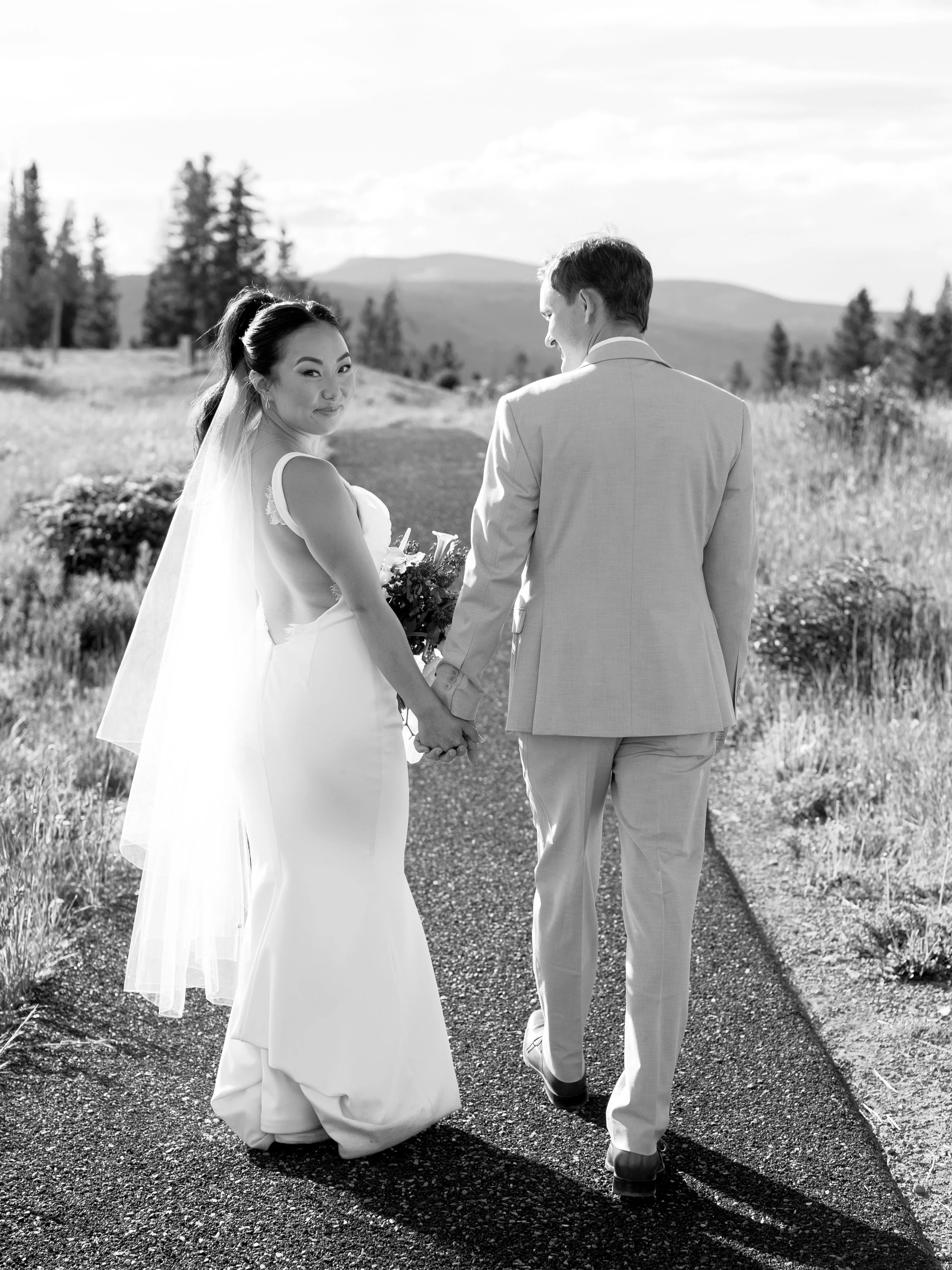 A bride and groom hold hands and walk down a path in a natural outdoor setting, both dressed in wedding attire, with trees and hills in the background.