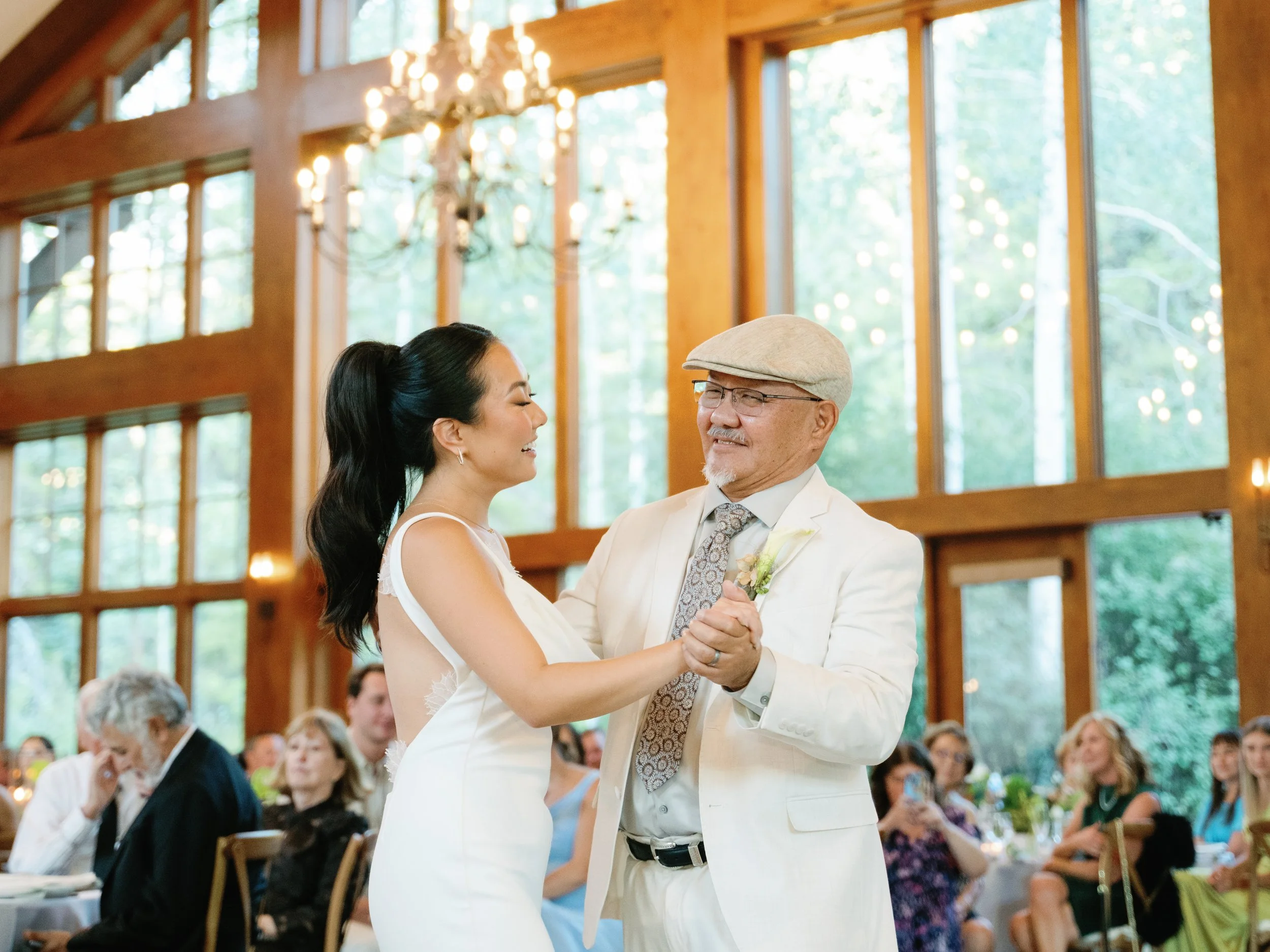 A bride and an older man, possibly her father, are dancing at a wedding reception in a rustic venue with large windows and wood paneling. The bride wears a white dress, and the man is dressed in a white suit with a beige cap. Guests are seated at tab