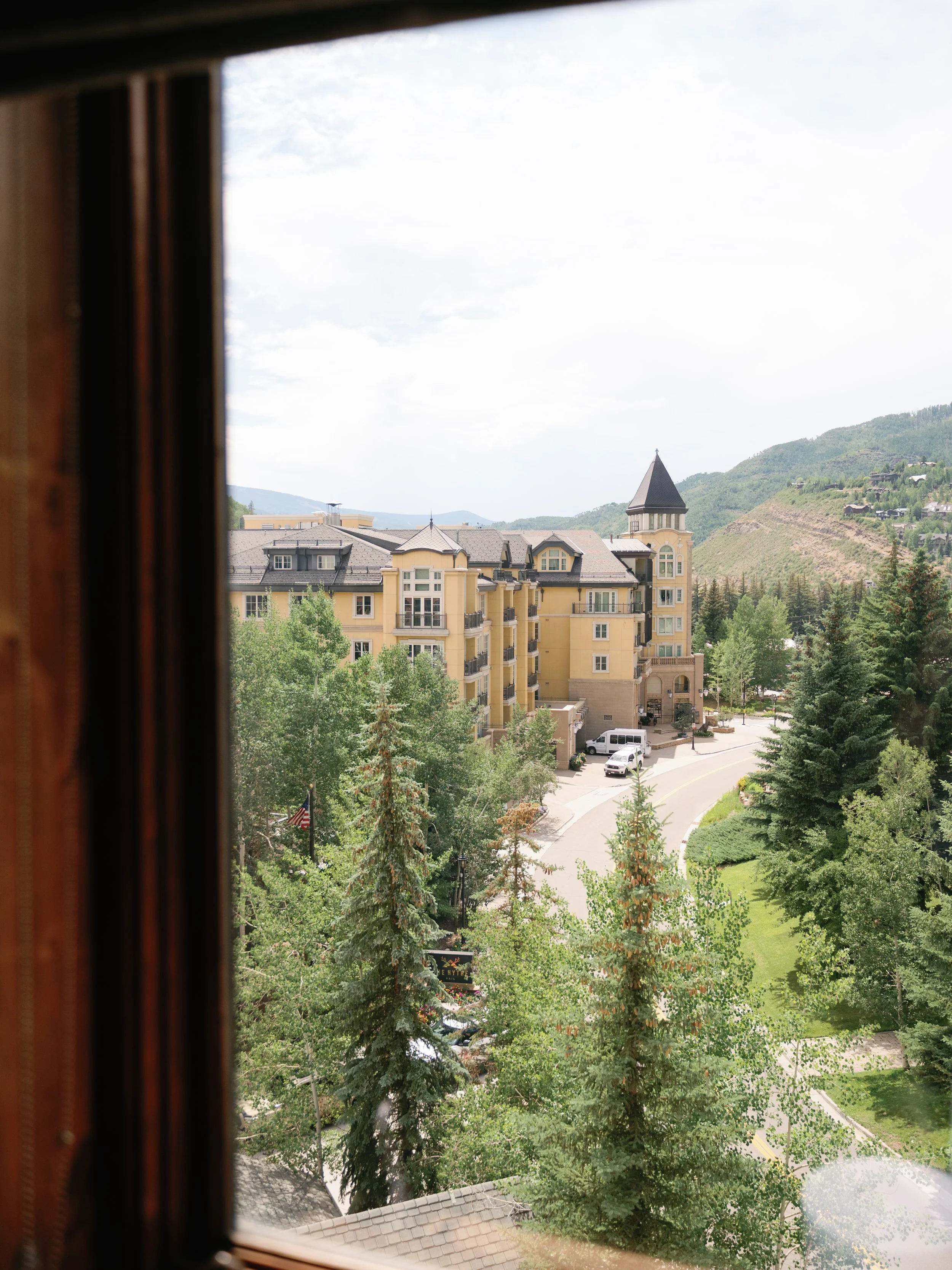 View through a window showing a large yellow residential building with a turret, surrounded by green trees, a road, and distant mountains.