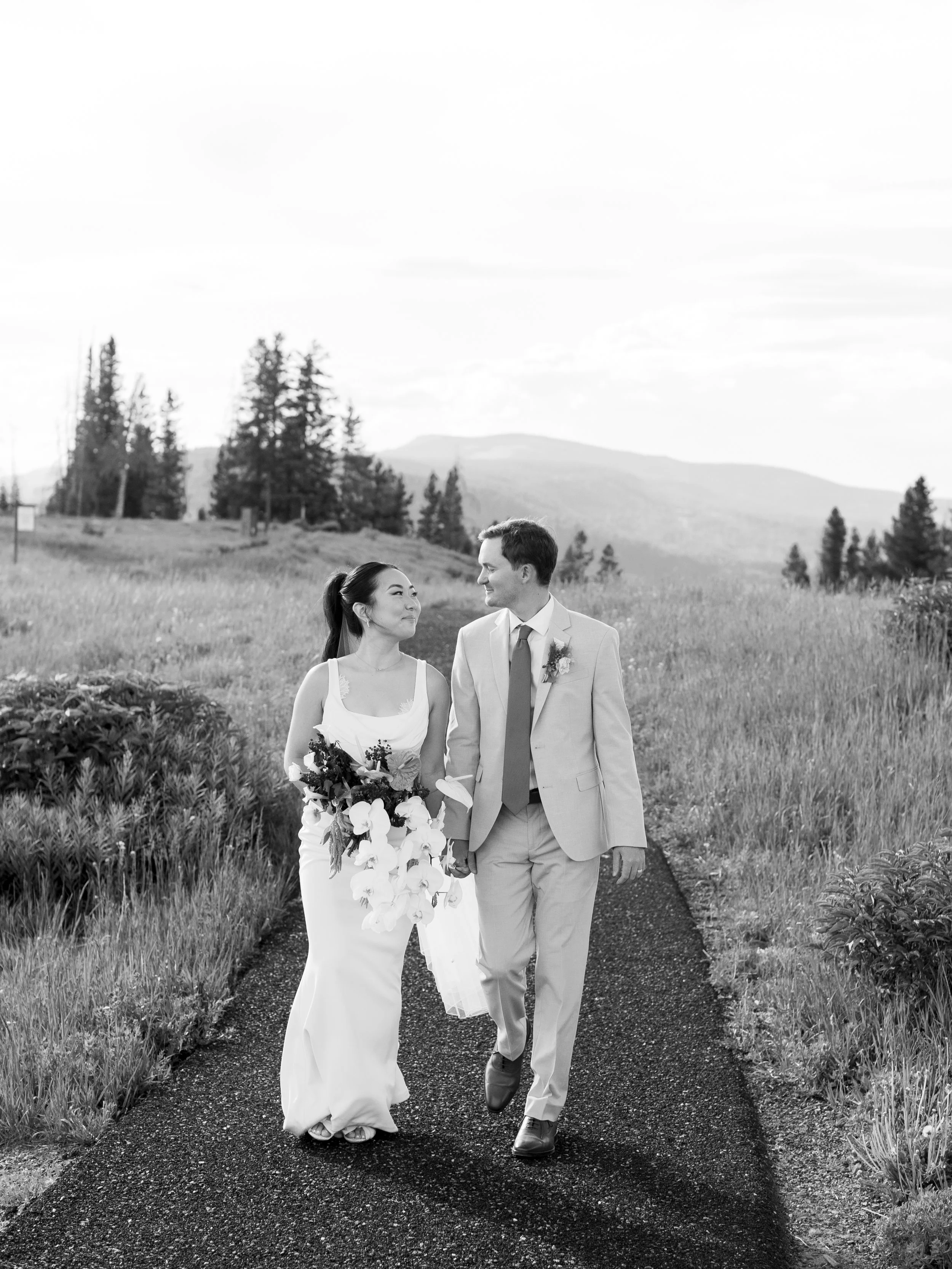 A black-and-white photograph of a newlywed couple walking on a rural path, holding hands, with trees and mountains in the background.
