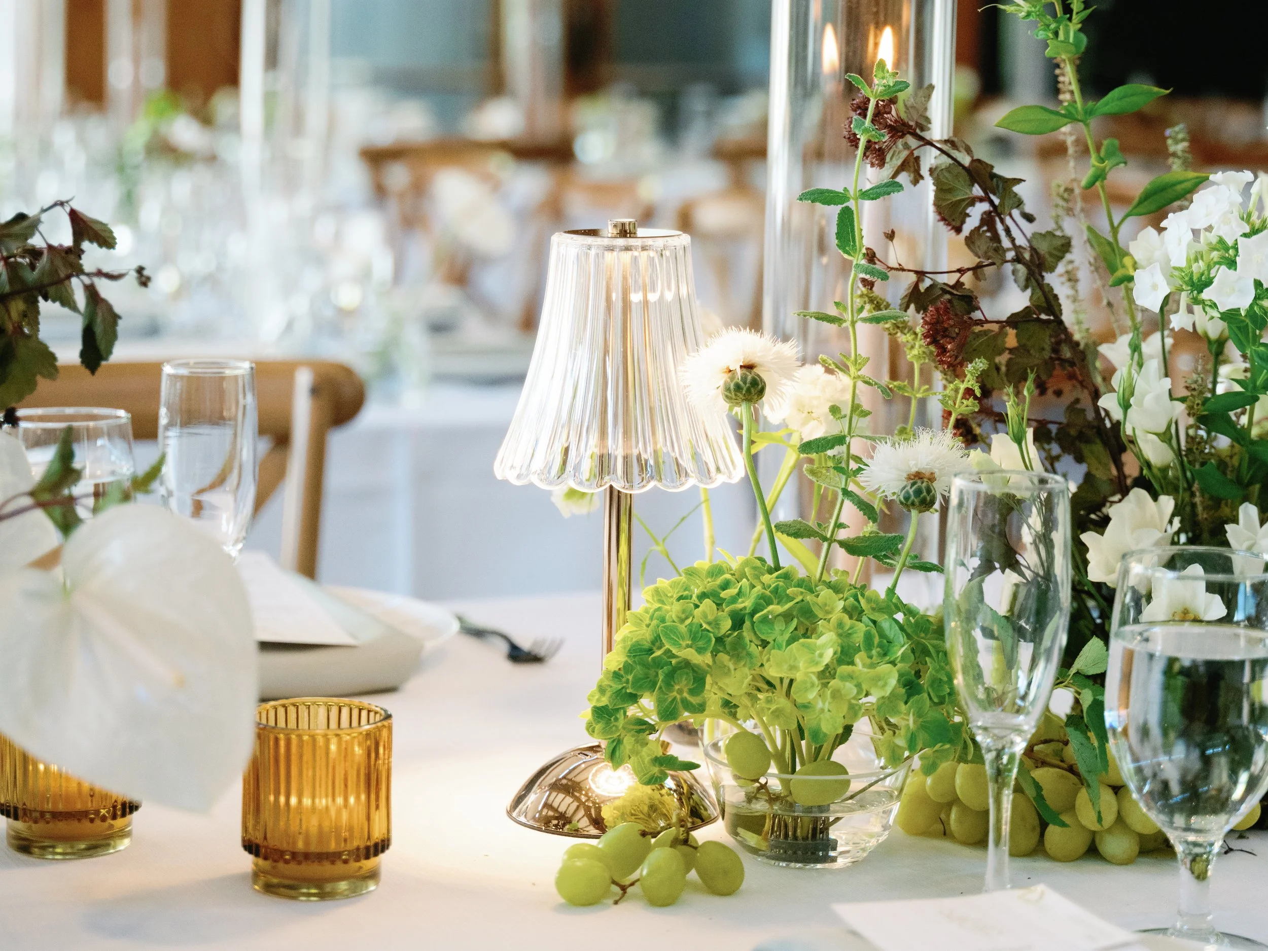 Elegant table setting with a floral centerpiece, glassware, gold candle holders, and a small table lamp with a glass shade.