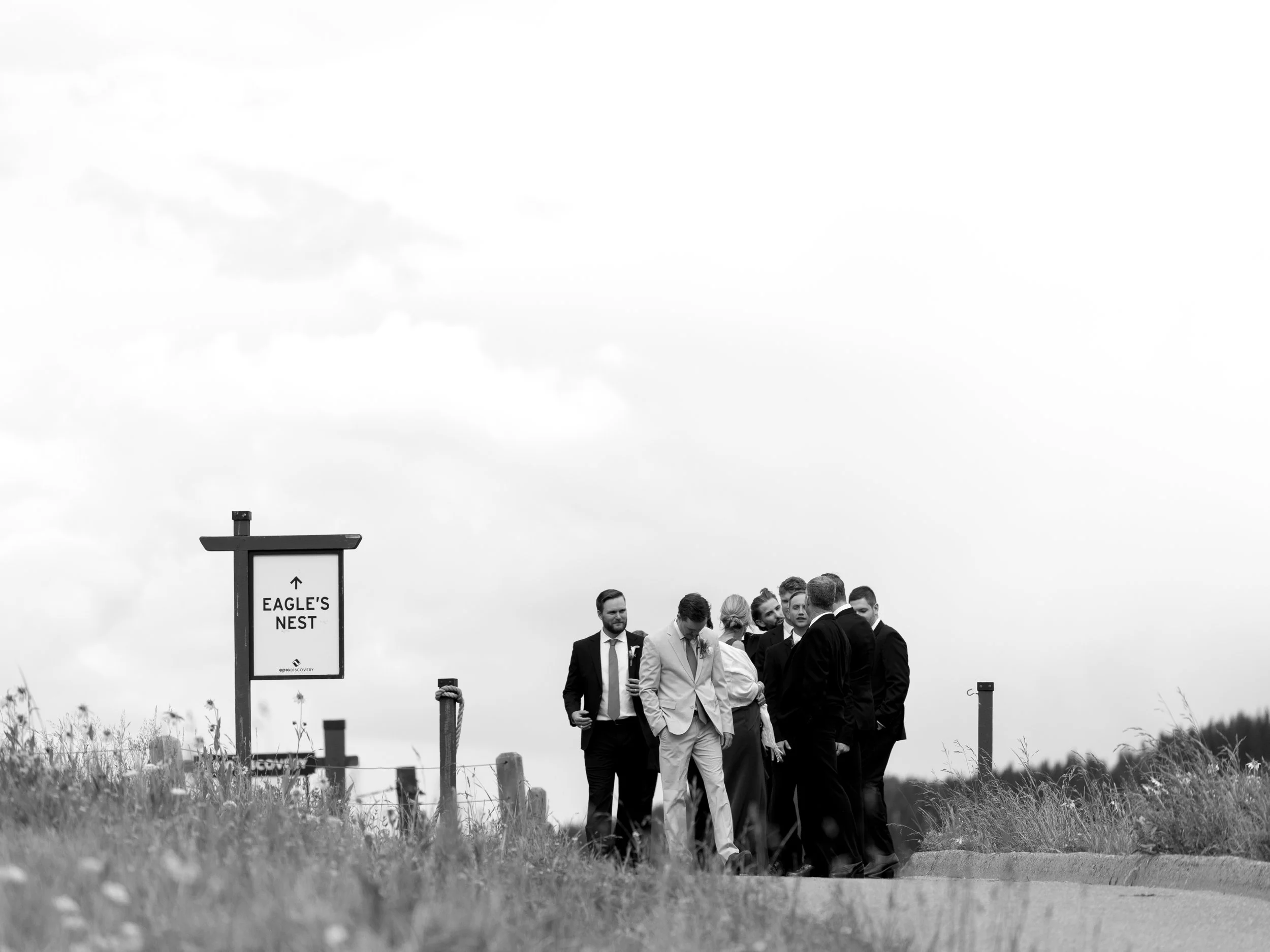 A group of people dressed in formal suits, standing on a street near a sign that reads "Eagle's Nest" with an arrow pointing upwards, in a rural outdoor setting with tall grass and cloudy sky.