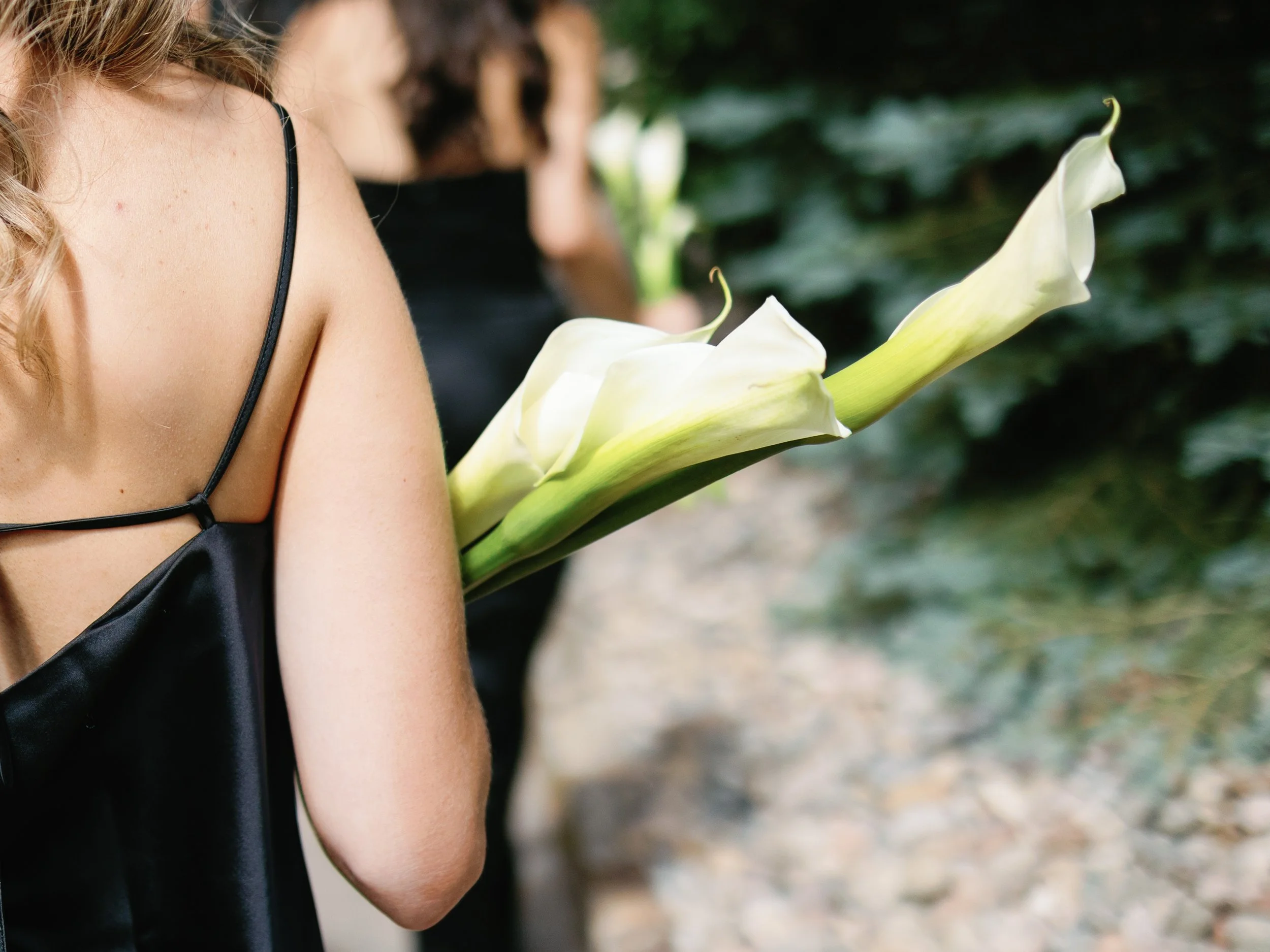 A woman wearing a black dress with spaghetti straps holding a white calla lily flower outdoors.