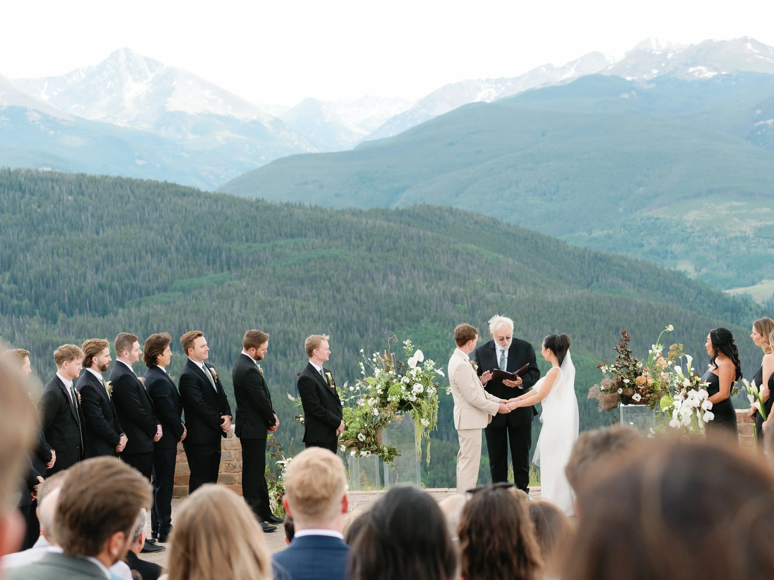 A wedding ceremony outdoors with mountains in the background. The bride and groom are holding hands, facing each other, with an officiant reading from a book. Bridesmaids and groomsmen are standing in a line behind the couple. Guests are seated and w