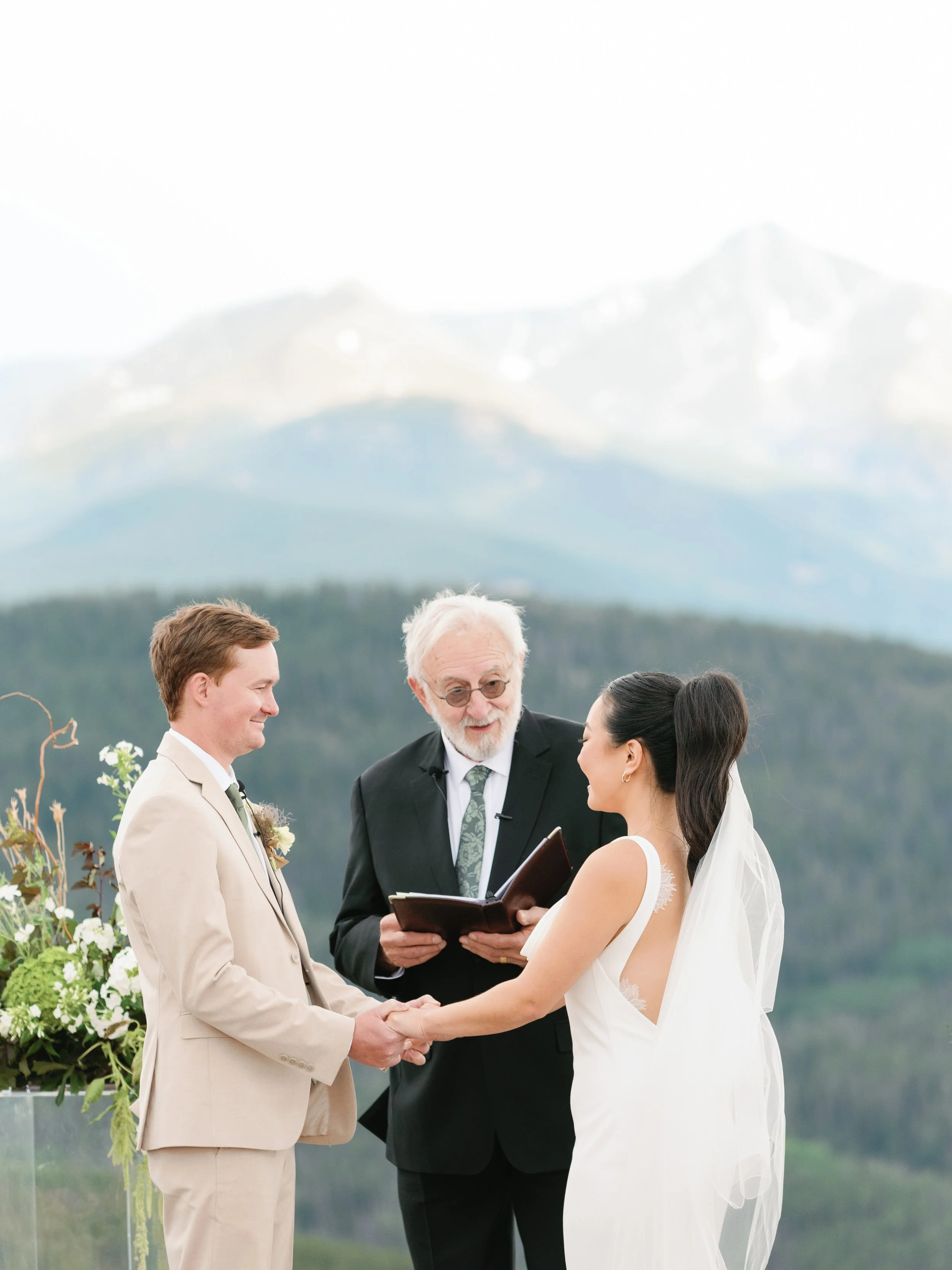 An outdoor wedding ceremony with a bride and groom holding hands, standing in front of an officiant during the vows with a mountain landscape in the background.