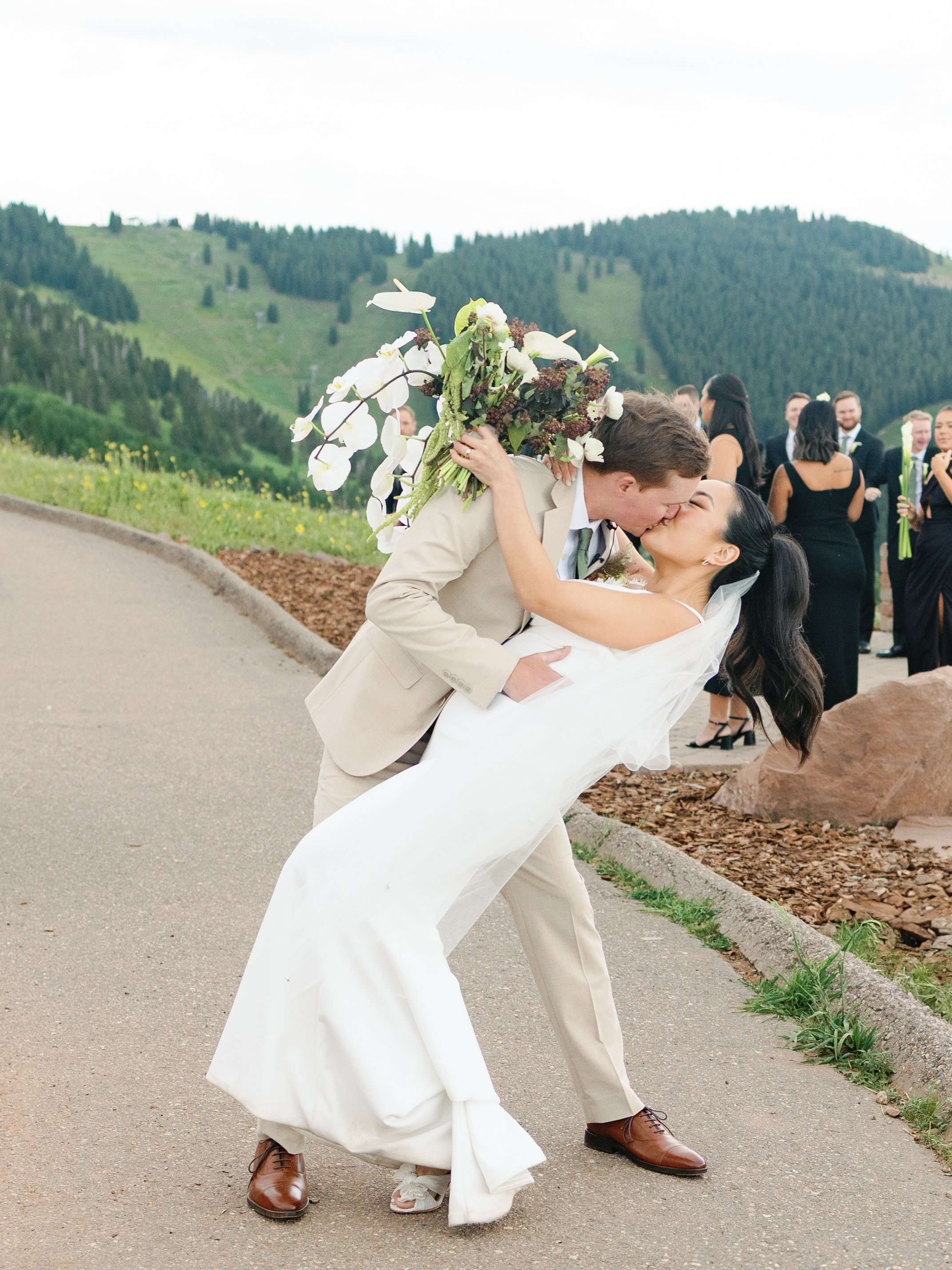 A newlywed couple sharing a kiss during their wedding celebration outdoors, with the groom in a beige suit and the bride in a white gown, holding a large bouquet of flowers, with a scenic mountain background and guests in the background.