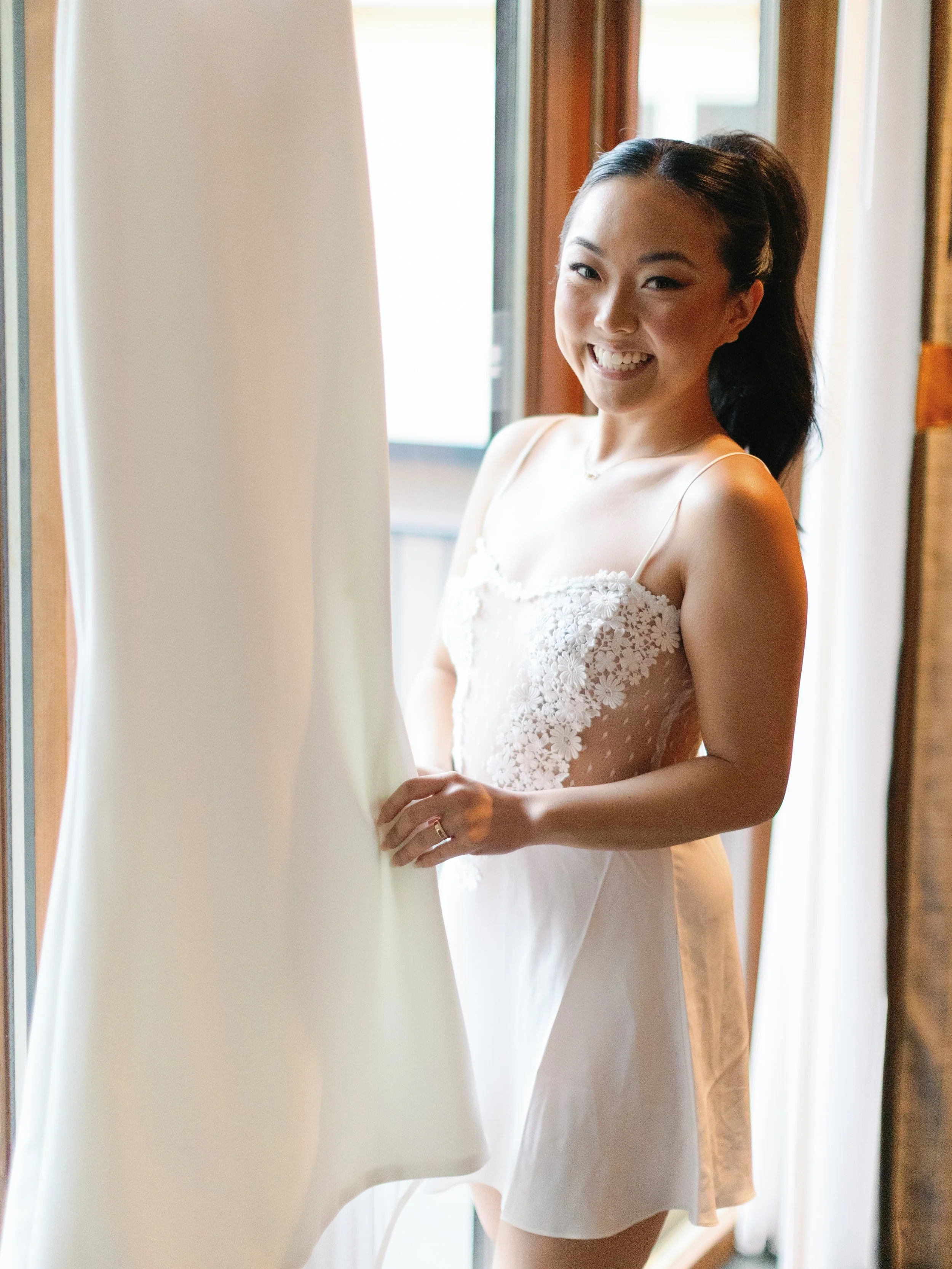 A young woman in a white dress with lace details is standing by a window, smiling and looking at the camera while holding a white fabric.