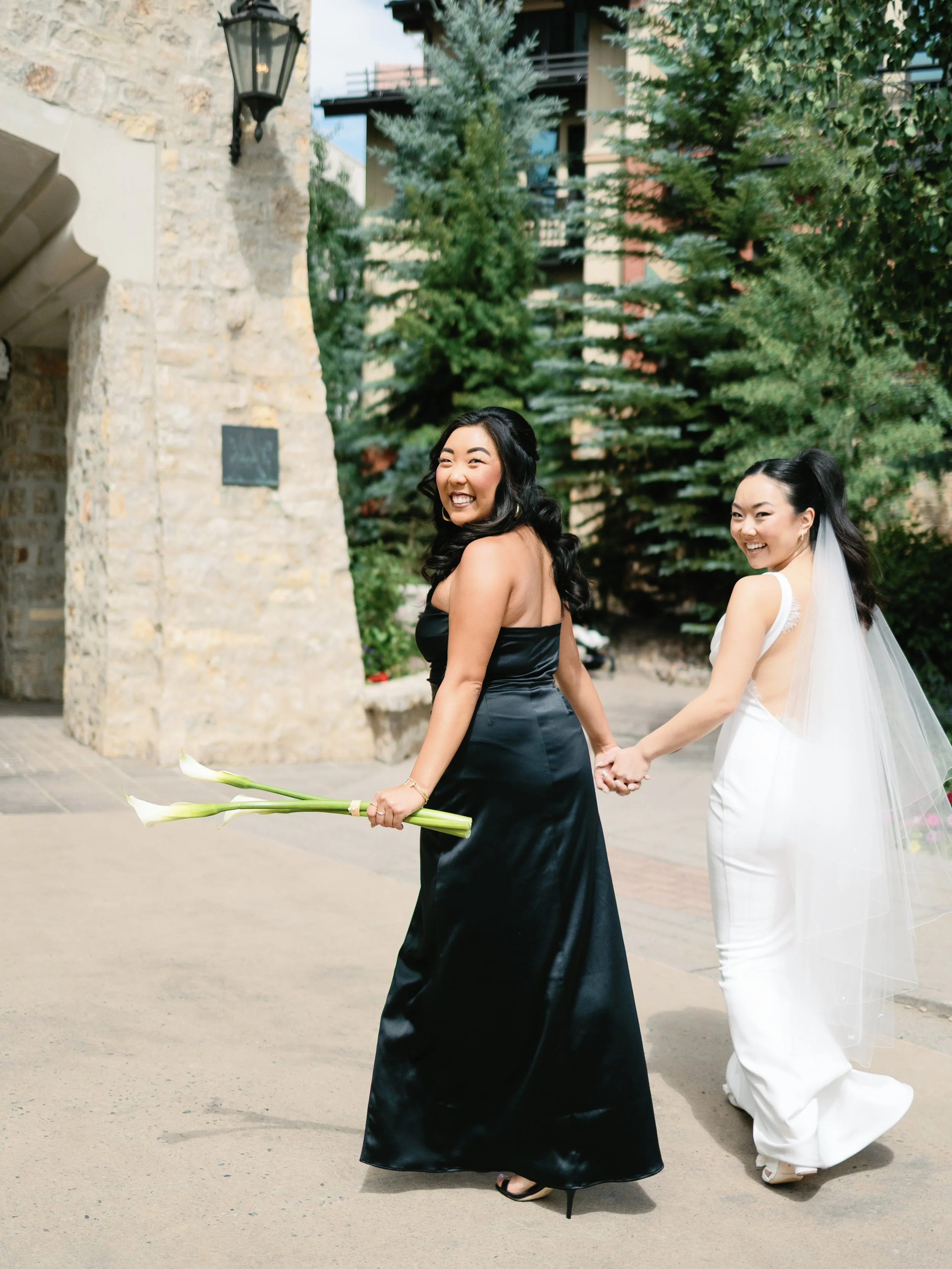 Two women, one in a black dress holding calla lilies, and one in a white wedding gown with a veil, holding hands and smiling, outdoors near a stone building and evergreen trees.