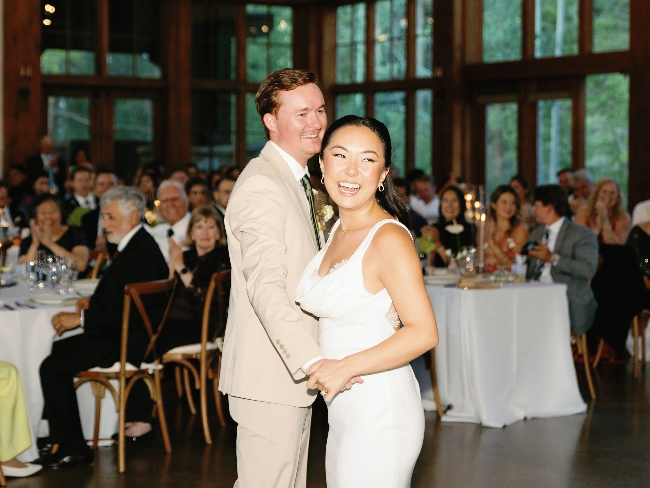 A smiling bride and groom dancing at their wedding reception, surrounded by seated guests in a wooden hall with large windows showing trees outside.