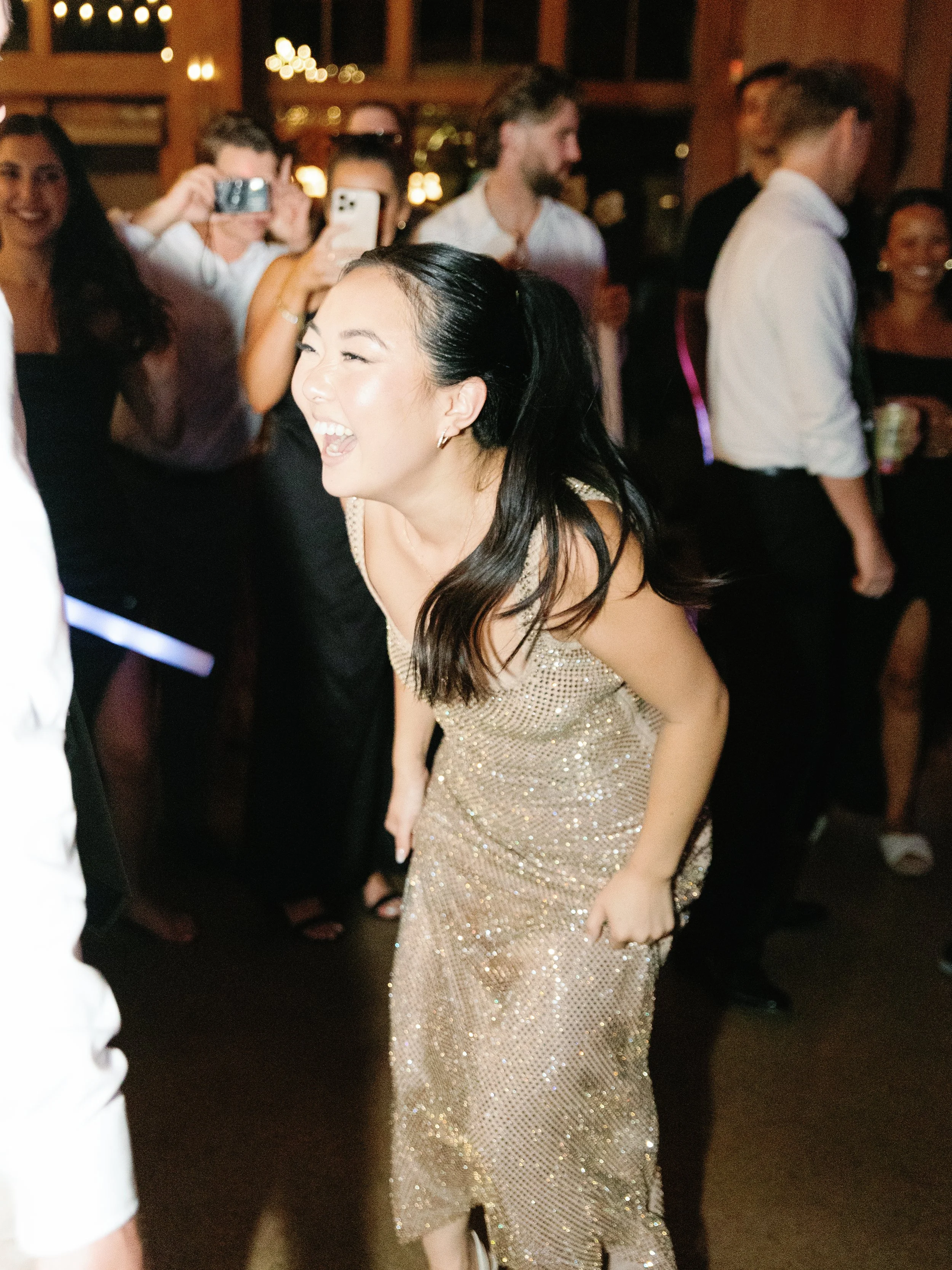 A woman in a gold sequin dress laughing and dancing at a lively party or celebration with other guests taking photos and socializing in a warmly lit indoor venue.