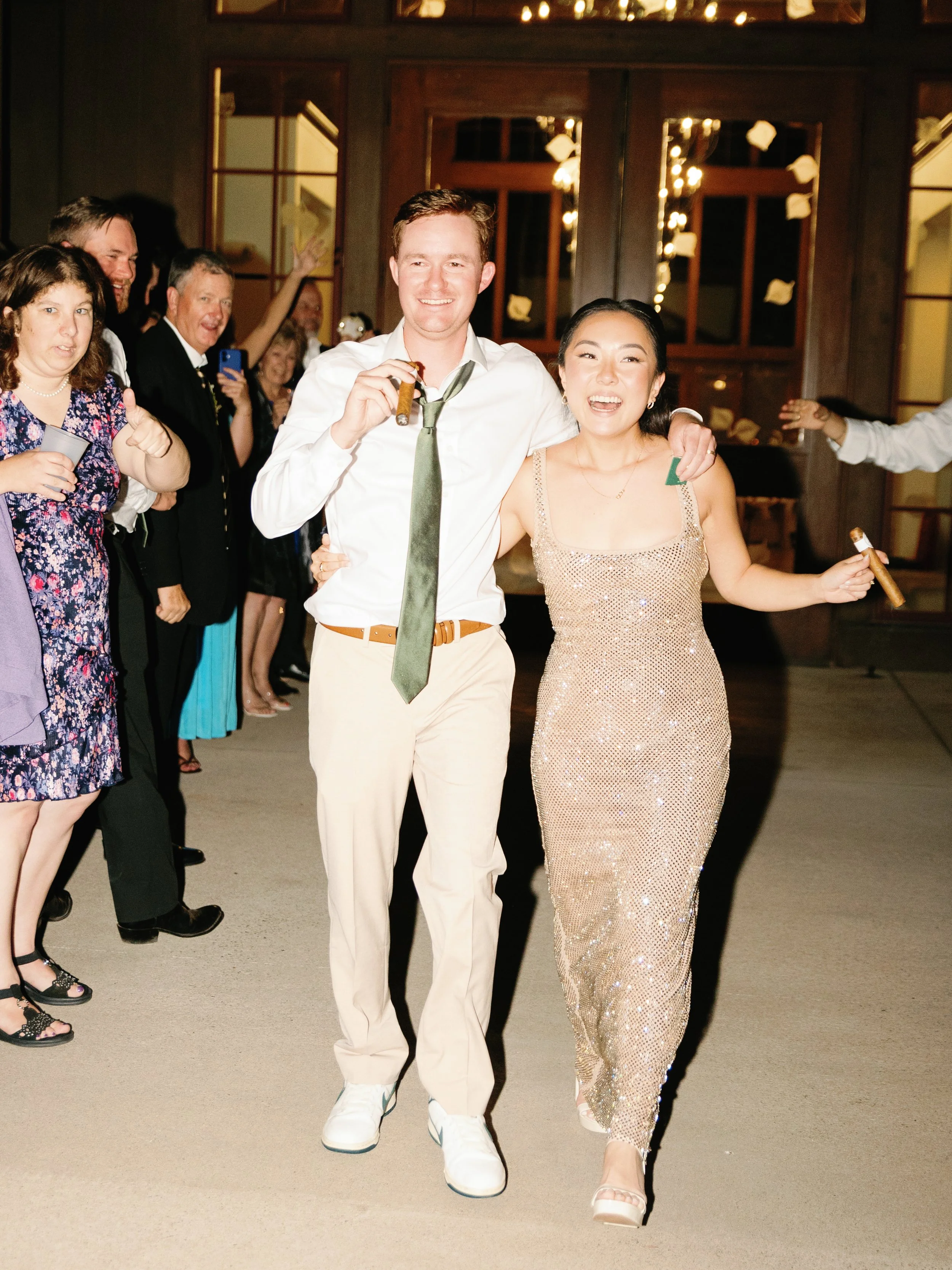 A joyful diverse group of wedding guests celebrating, with two central people smiling and holding cigars, surrounded by friends in formal attire, indoors with warm lighting and wooden decor.