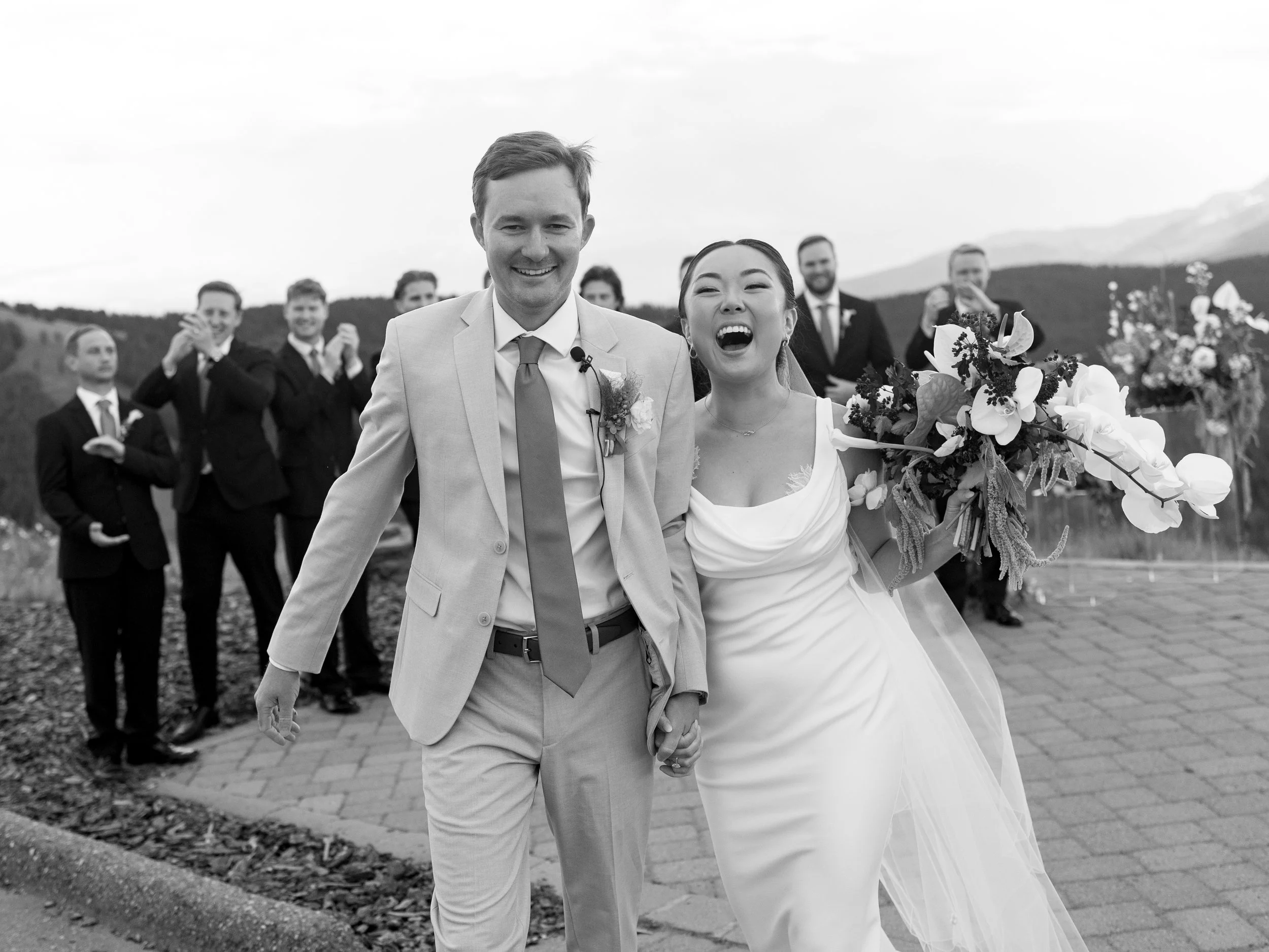A happy bride and groom walking arm in arm during their wedding reception or photoshoot, holding a bouquet of flowers, with groomsmen and bridesmaids in the background, outdoors in a scenic location.