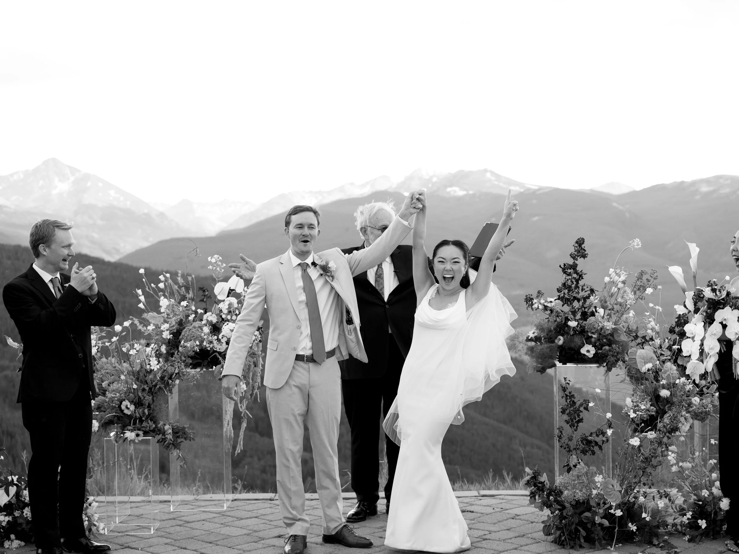 A joyful wedding celebration with a bride and groom holding hands and raising their arms, surrounded by friends, with mountains in the background.