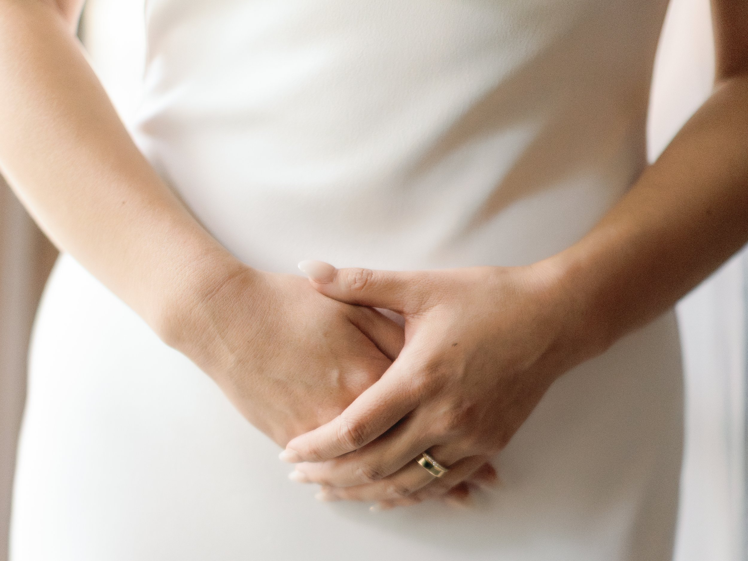 Close-up of a person’s hands gently holding their pregnant belly, wearing a wedding ring, dressed in white clothing.