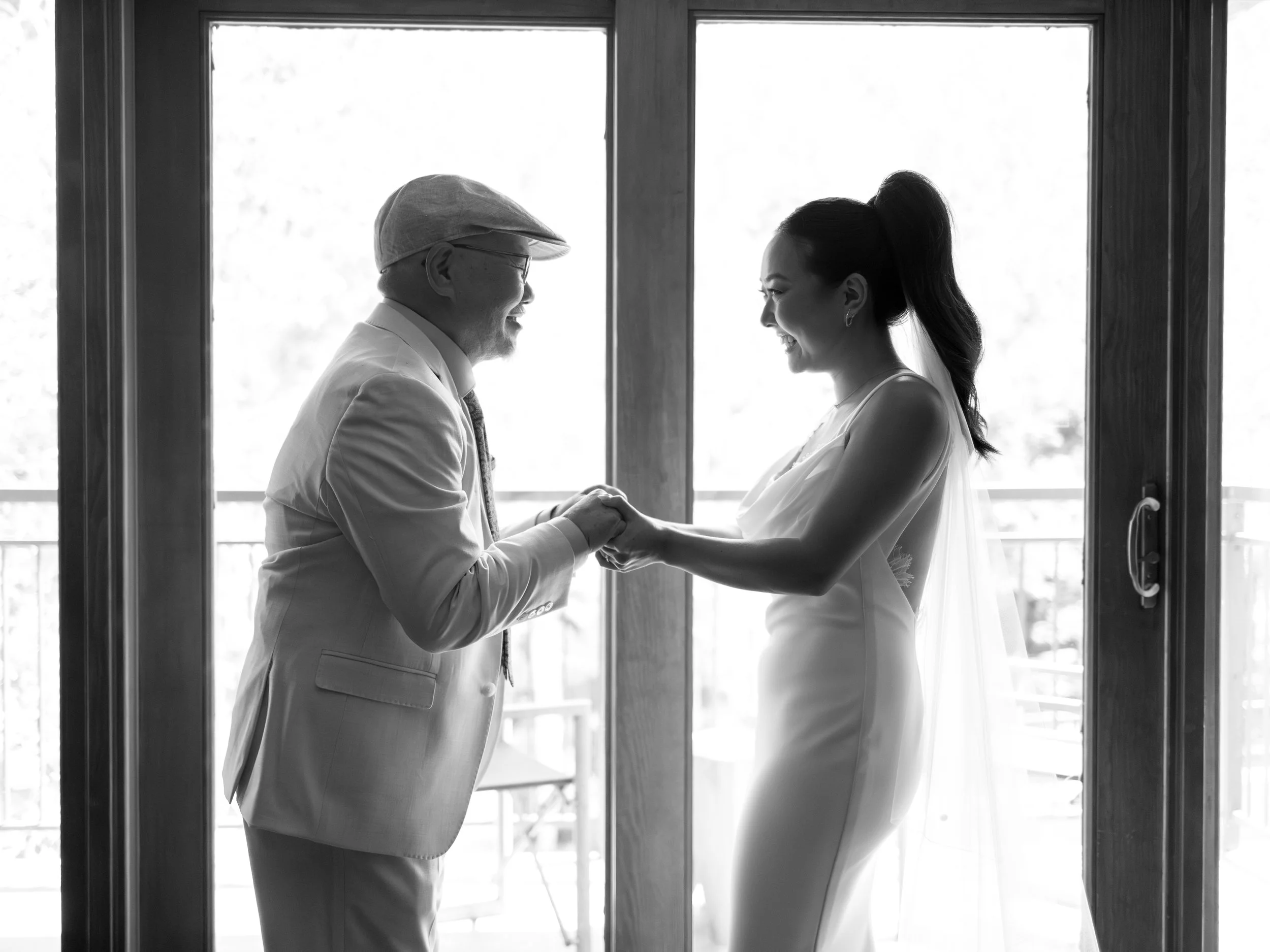 A bride and an older man holding hands and smiling in front of glass doors.