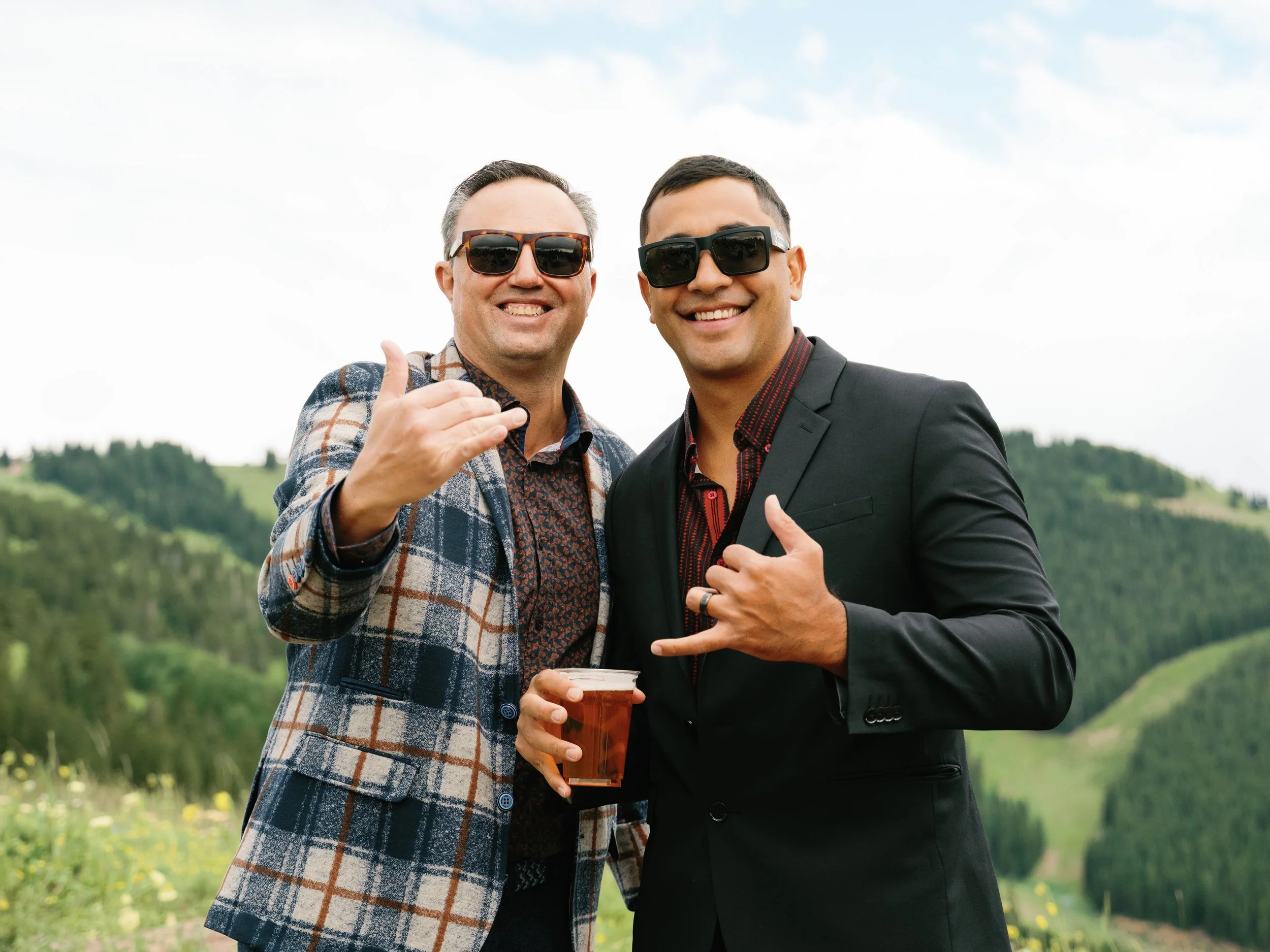 Two men in sunglasses smiling outdoors, making hand gestures, holding drinks, with a scenic green landscape and hills in the background.