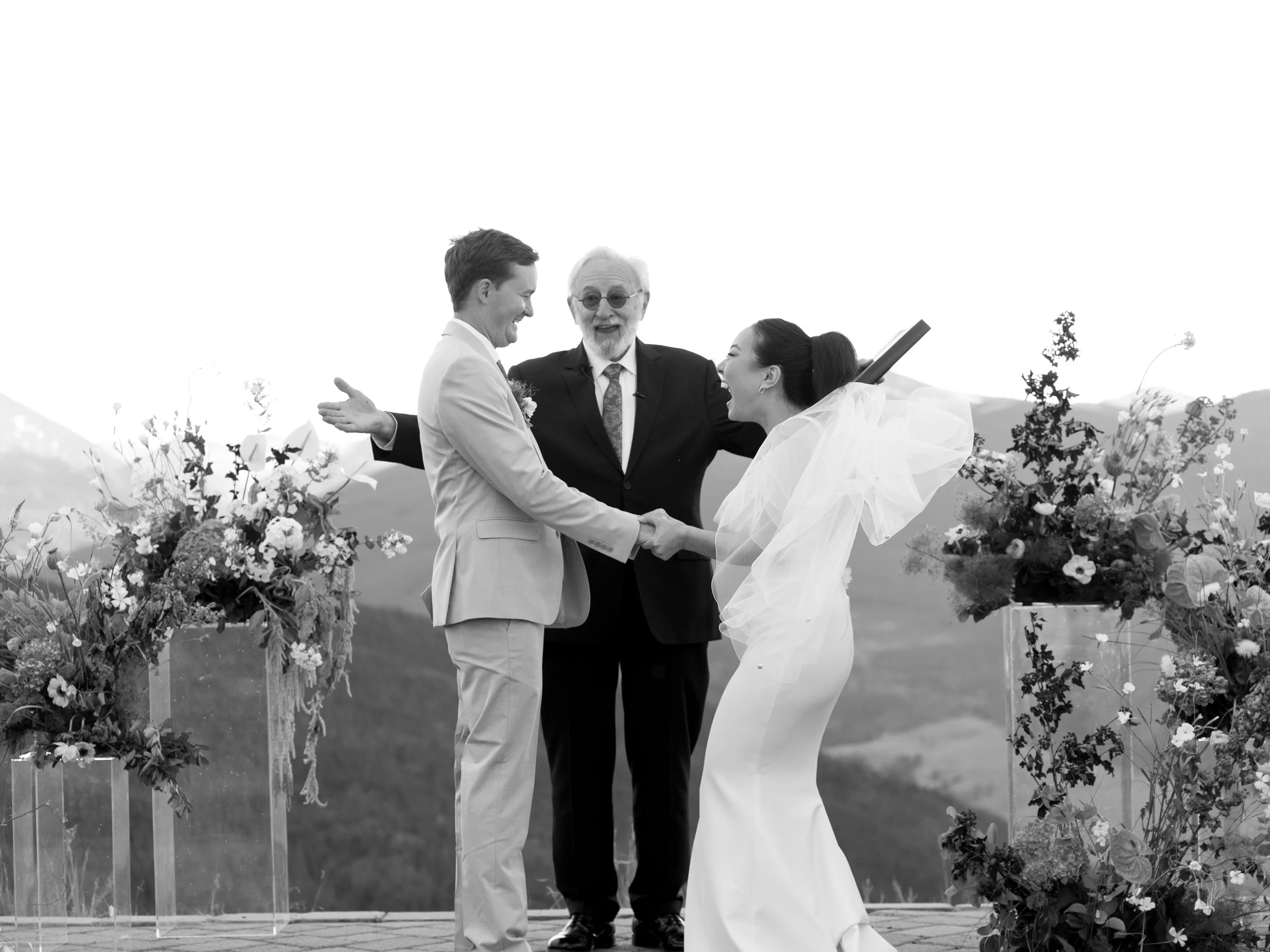 A couple's wedding ceremony outdoors with an officiant, holding hands and facing each other, surrounded by floral arrangements, with mountains in the background.