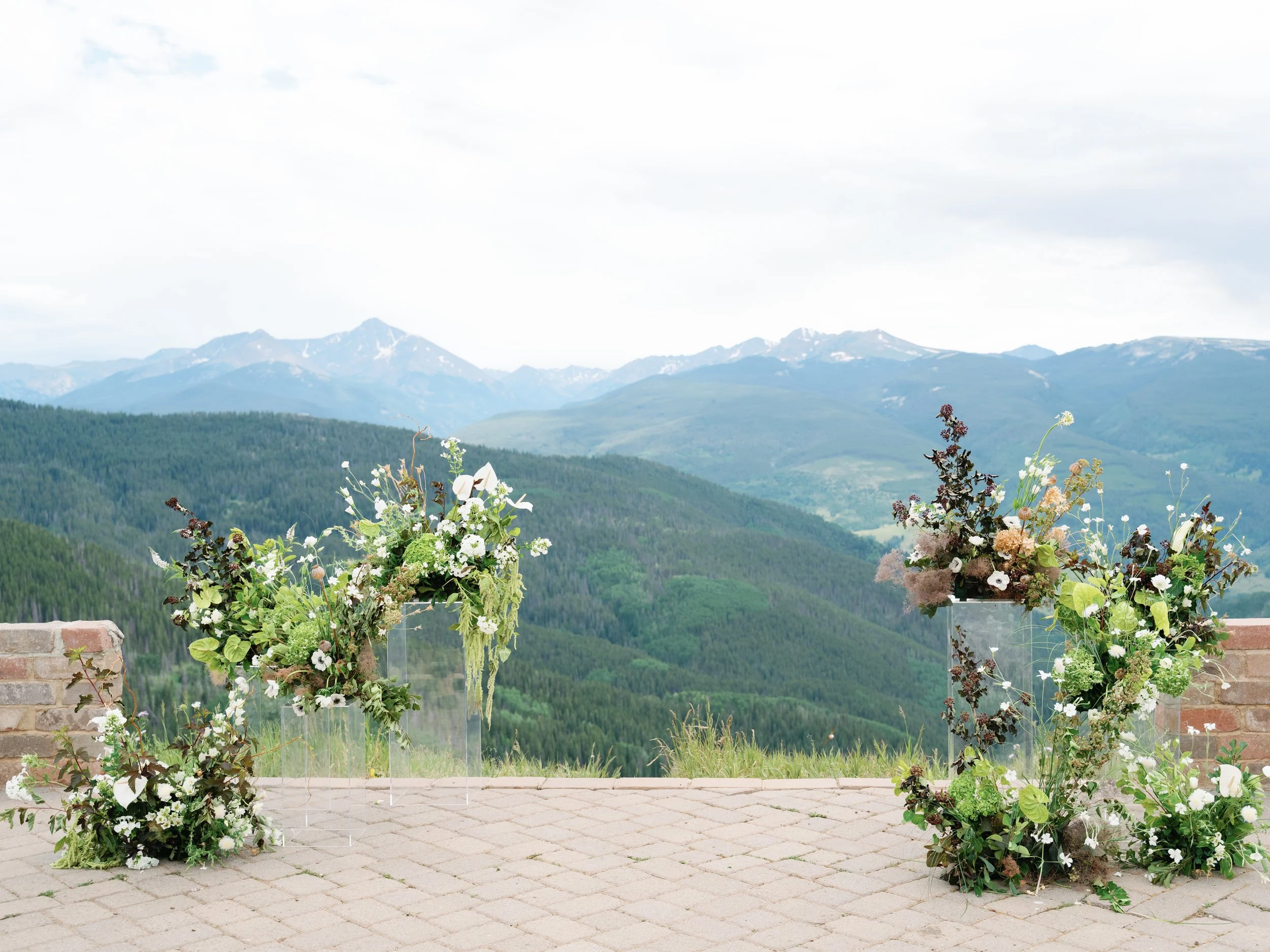 A scenic outdoor view of a mountain landscape with green hills and snow-capped peaks in the background, with three floral arrangements on a paved surface in the foreground.